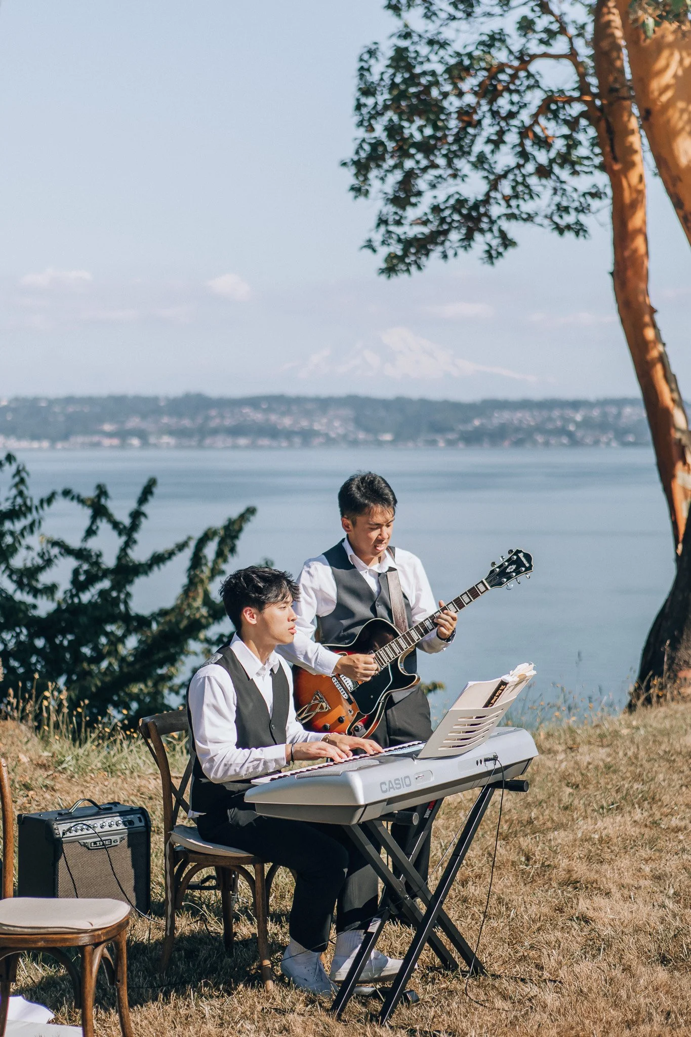 Two musicians playing keyboard and acoustic guitar beside a lake.