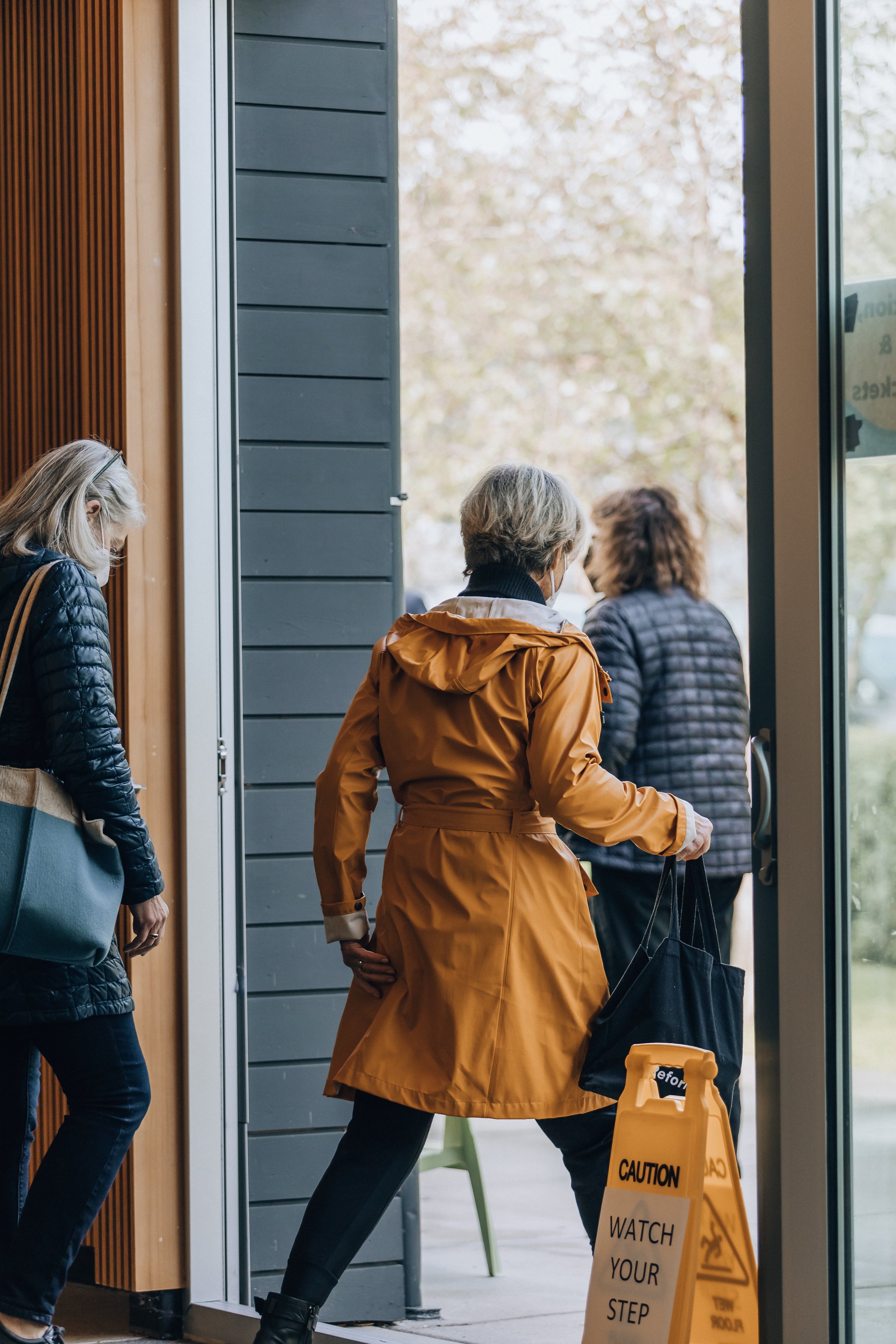 People entering a building, one with a yellow caution sign on the floor reading 'Watch Your Step' and a person holding a black bag, while the others are wearing jackets and masks.