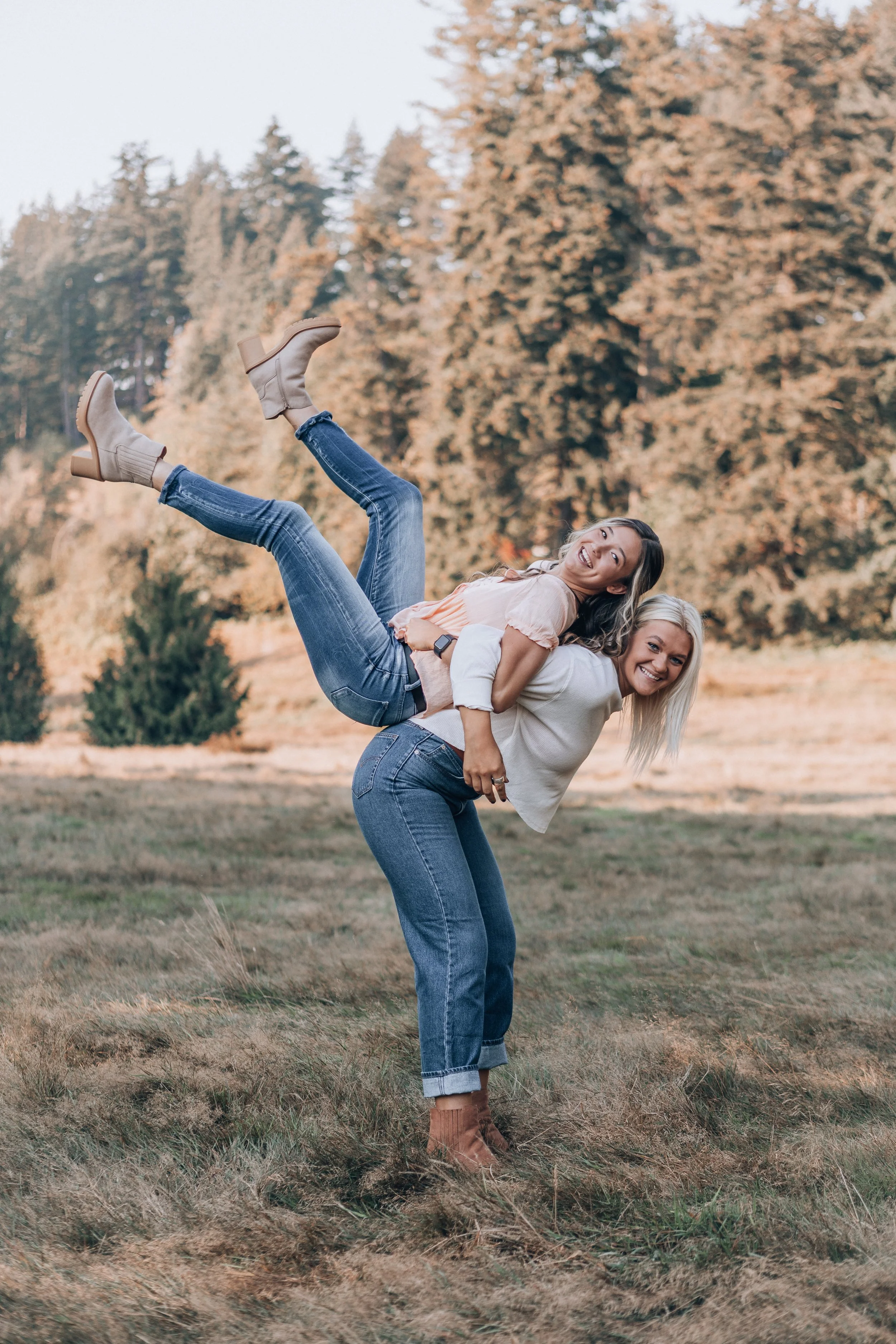 Two women enjoy a playful moment outdoors in an open field with autumn trees in the background. One woman stands and supports the other, who is lying across her shoulders with her legs extended upward. Both women are smiling and dressed casually.