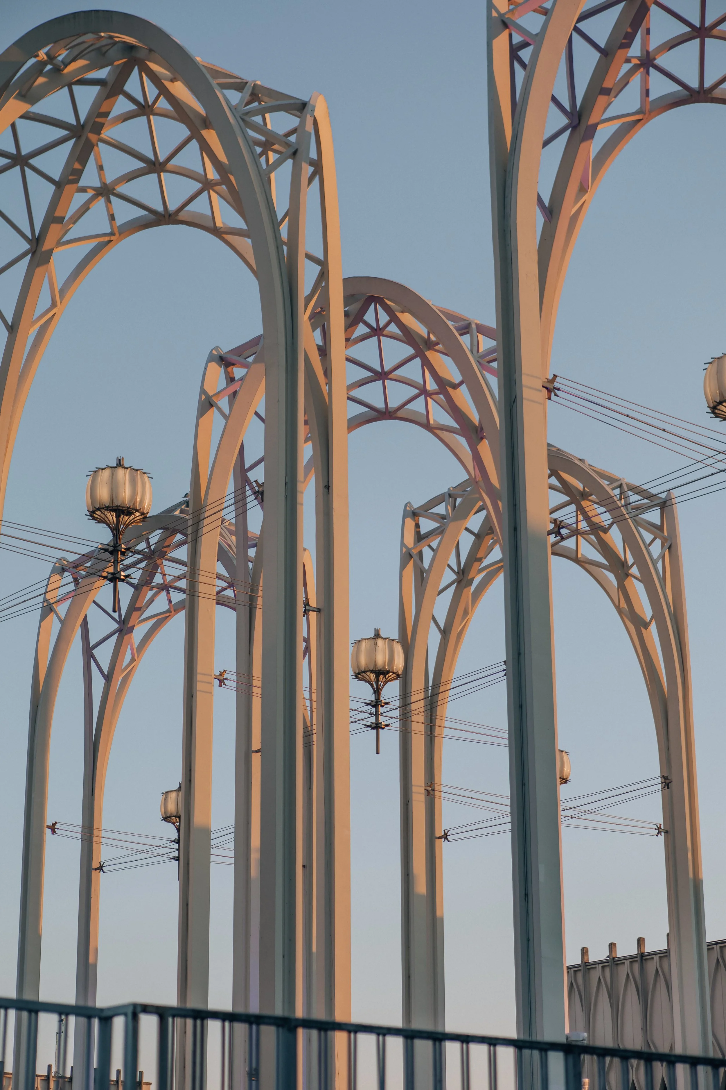 Close-up view of a Ferris wheel structure with white arches and support beams, under a clear blue sky, with some decorative lamps and wires visible.
