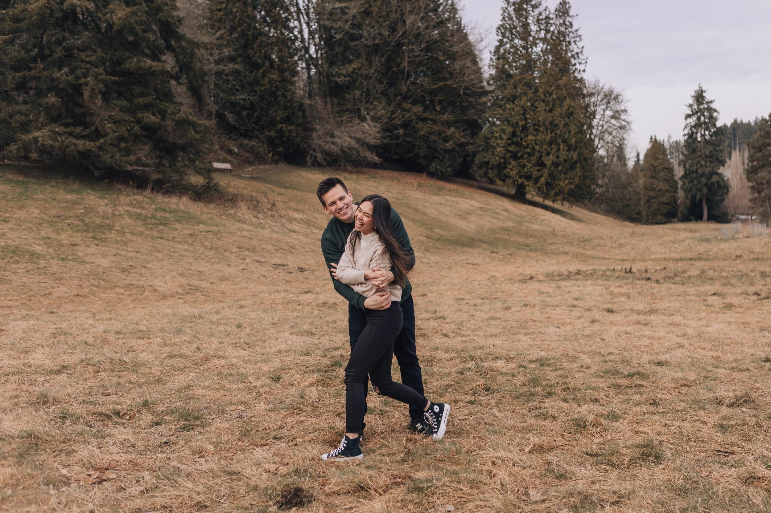 A young couple enjoying a playful moment outdoors in a park, with the woman being lifted from behind by the man, both smiling and laughing. The park has grass and large trees in the background on an overcast day.