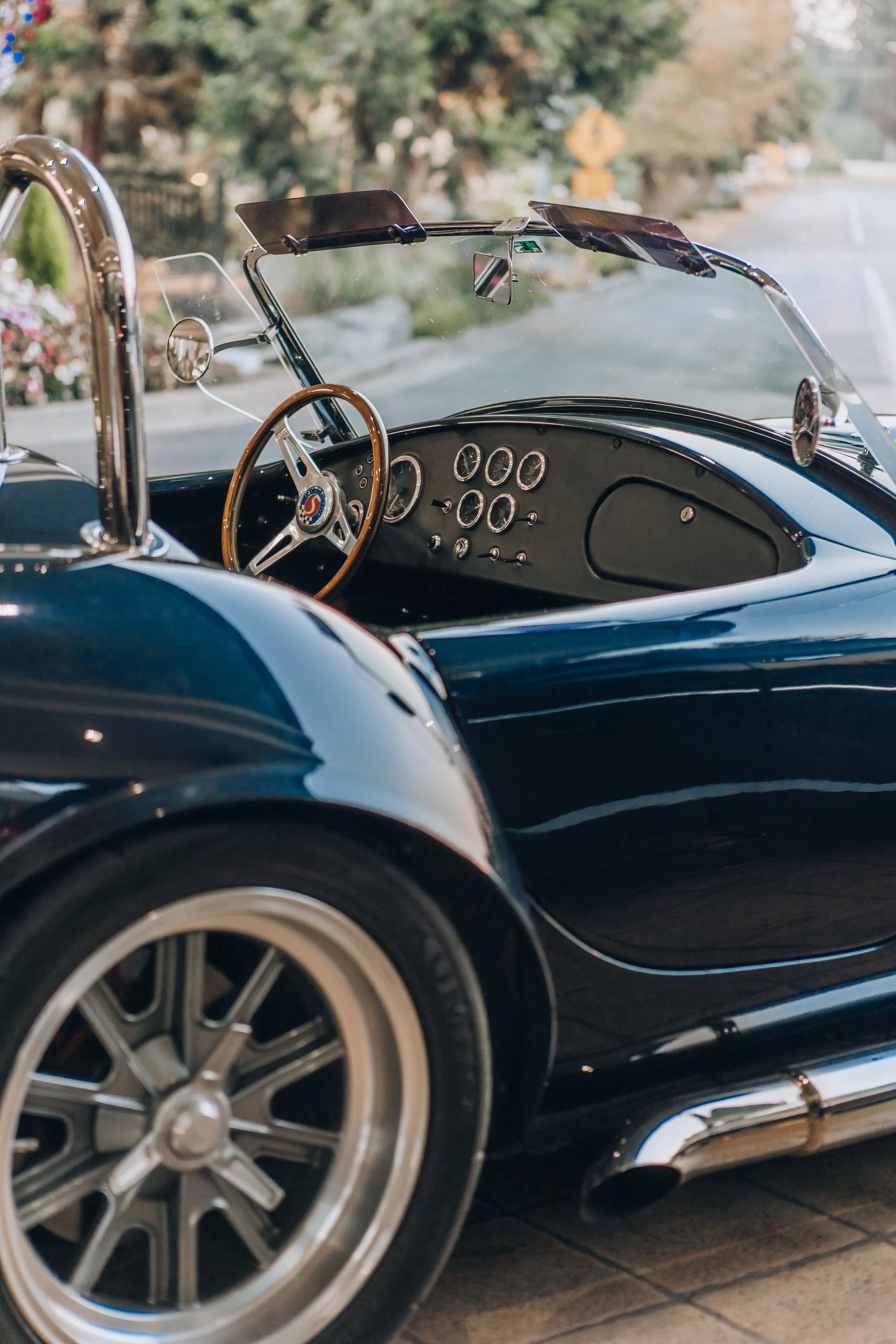 Close-up of the interior of a classic car with a wooden steering wheel, dashboard gauges, and a clear windshield, parked on a street with trees and sidewalk in the background.