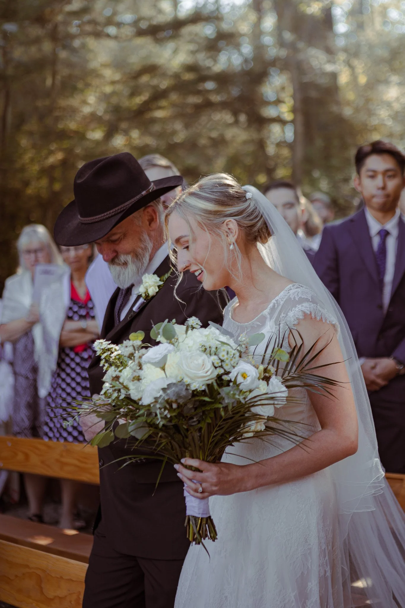 A bride holding white flowers beside a man in a dark suit and hat.