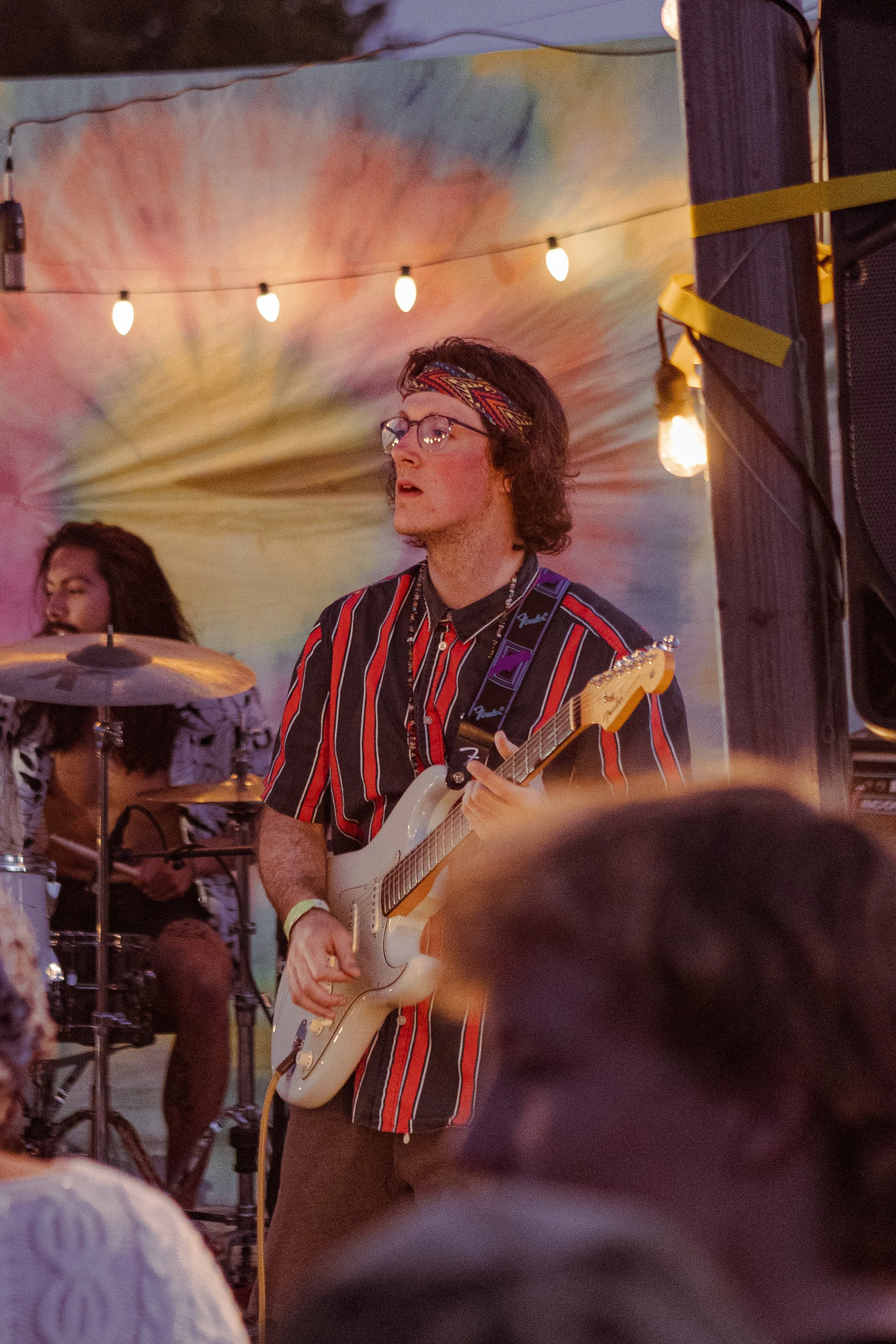 A person with glasses, a colorful patterned headband, and a striped shirt playing an electric guitar during a live outdoor music performance at dusk. There are string lights above, and a person with long hair appears in the background, possibly singing or playing an instrument.