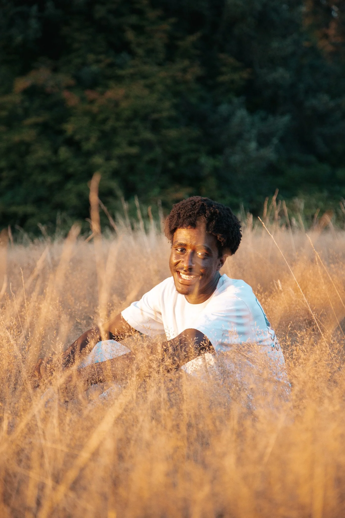 A young man with dark skin and curly hair, smiling, sitting in a field of tall grass during sunset or sunrise.