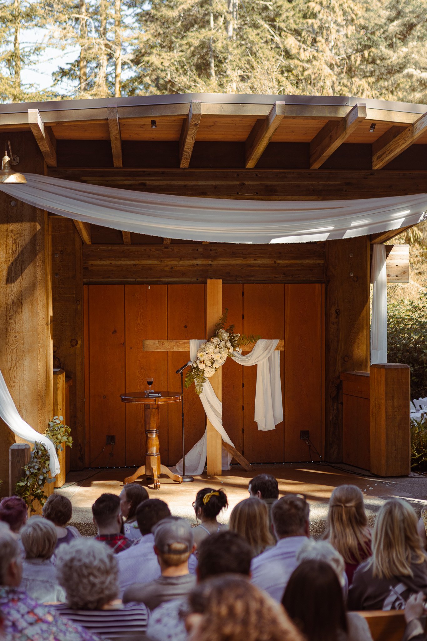 Seated wedding guests facing white drapery and flowers beneath a wooden pavilion.
