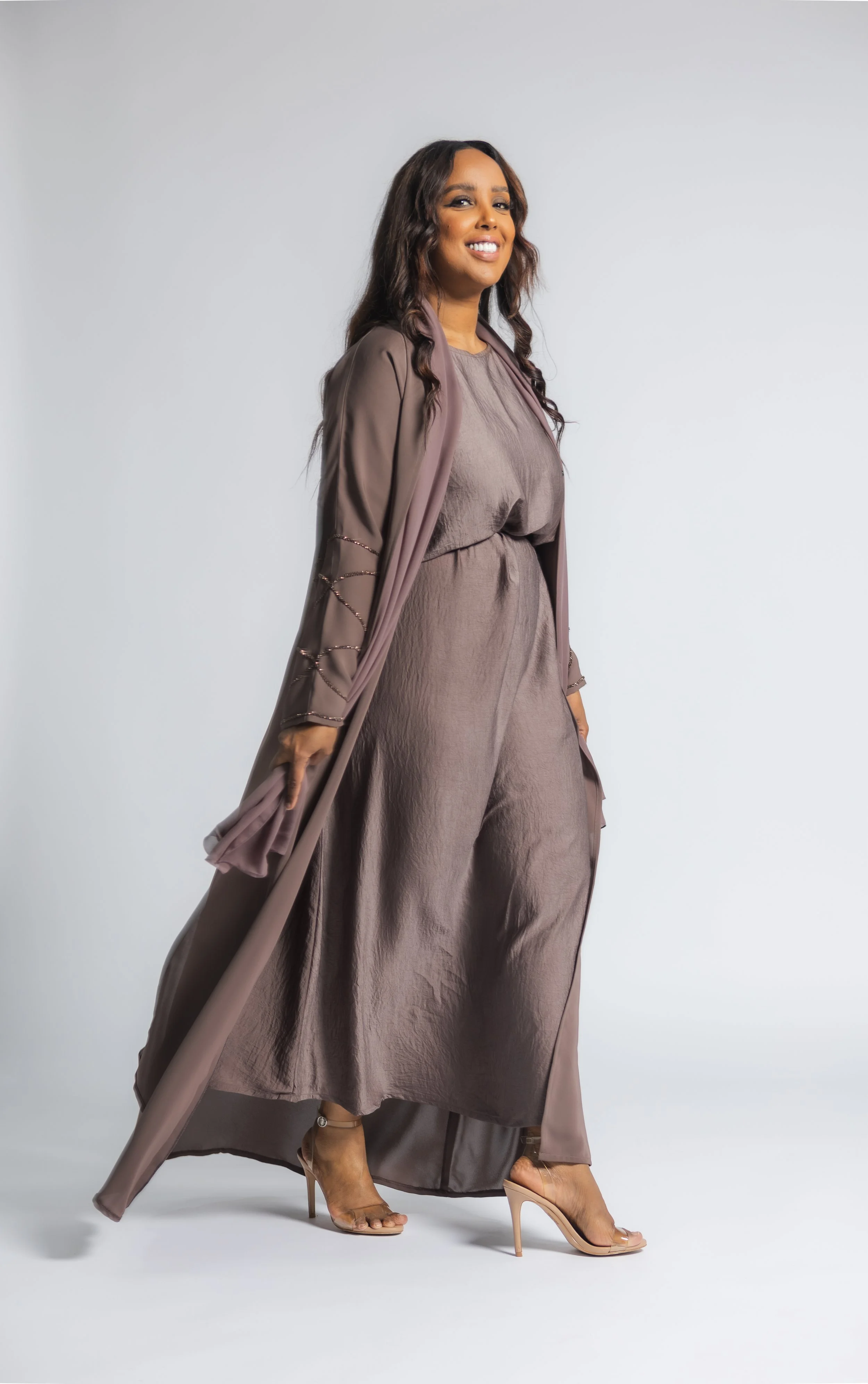 A woman in a taupe dress and sheer robe with embellishments, wearing nude high heels, smiling and posing against a plain light gray background.