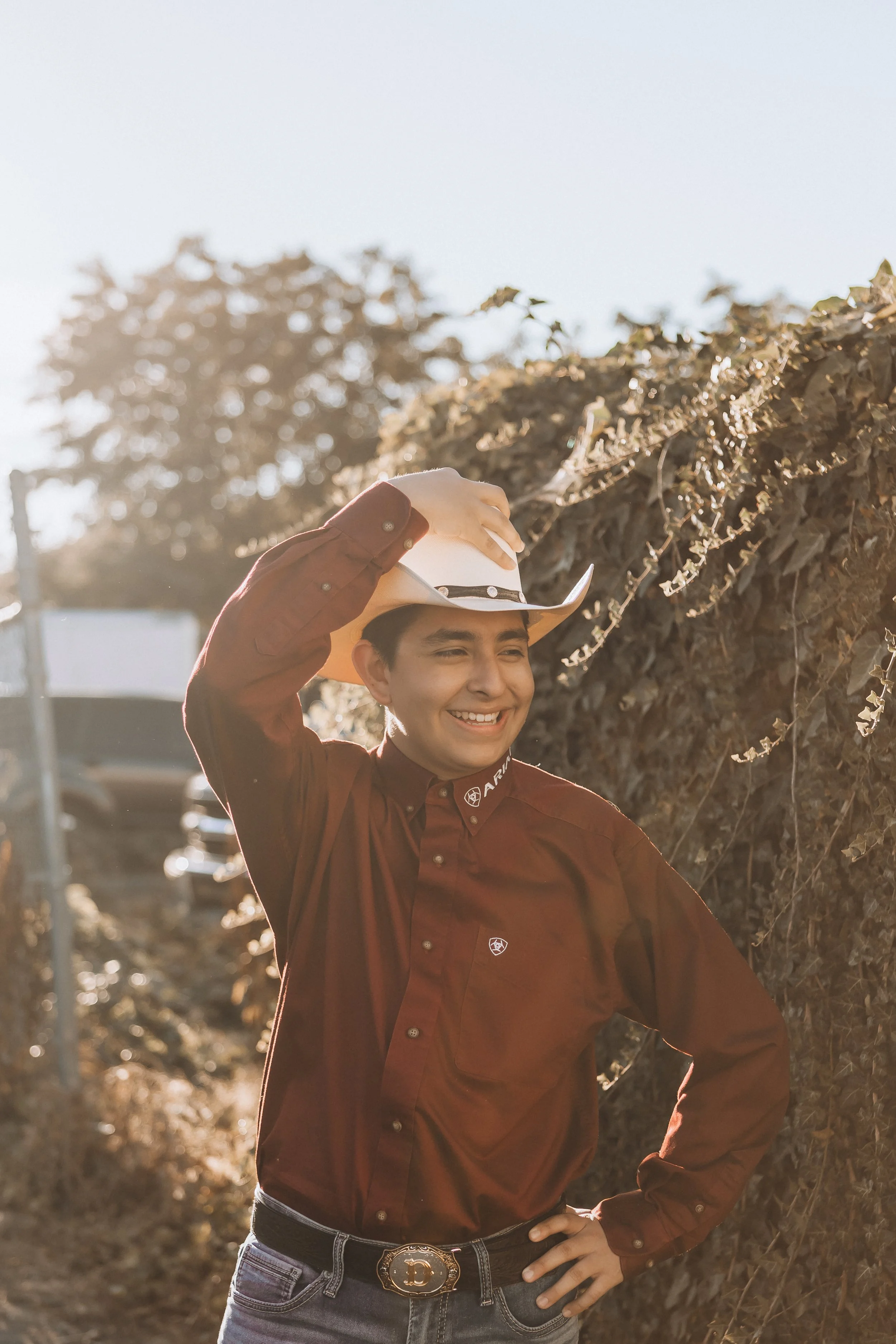 A young woman dressed as a cowboy, smiling and adjusting her white cowboy hat with her right hand, wearing a maroon plaid shirt and jeans with a decorative belt buckle, standing outdoors in sunlight near a hedge and trees.