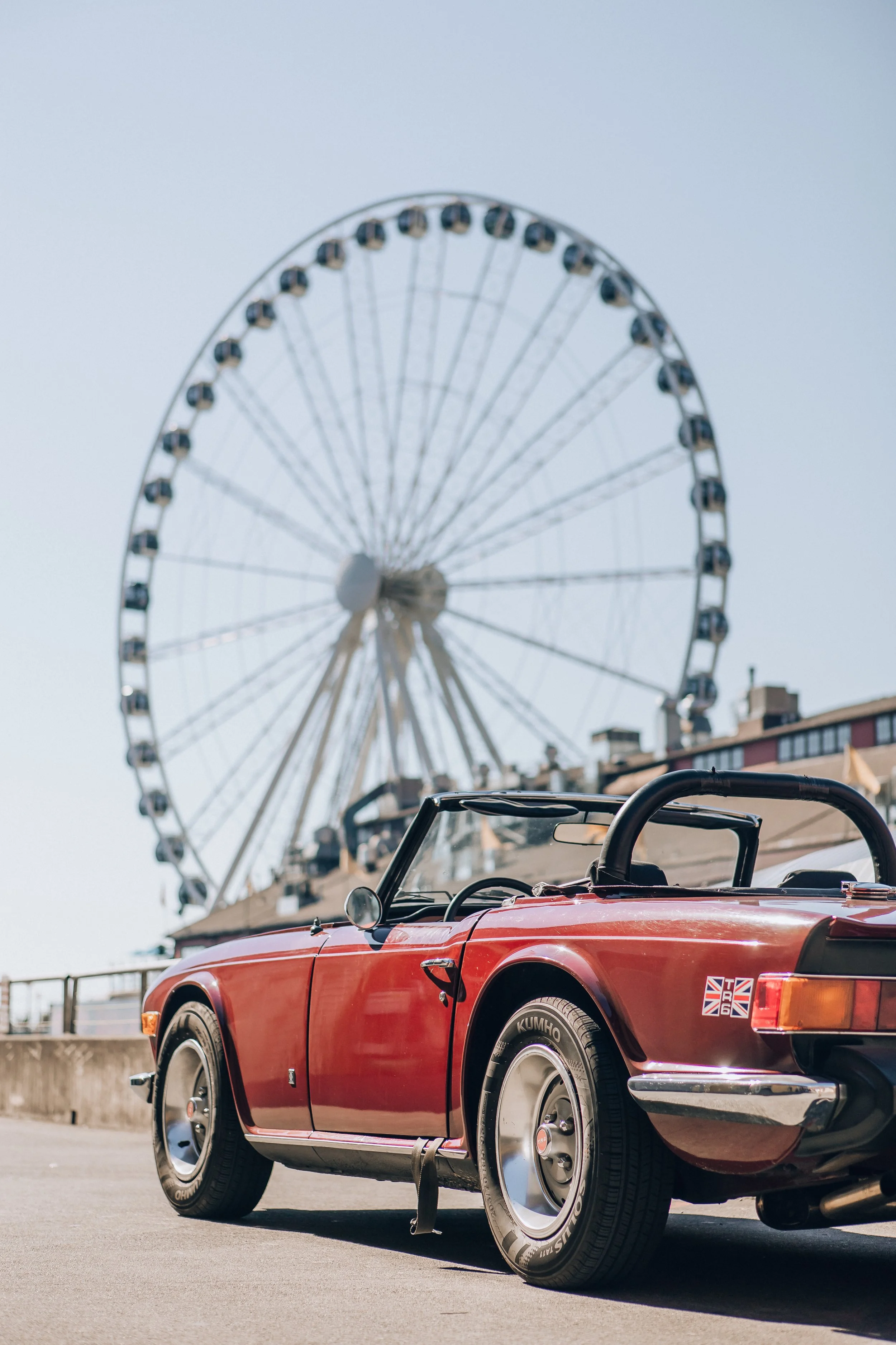 Classic red convertible car parked near a Ferris wheel at an outdoor fair or amusement park.