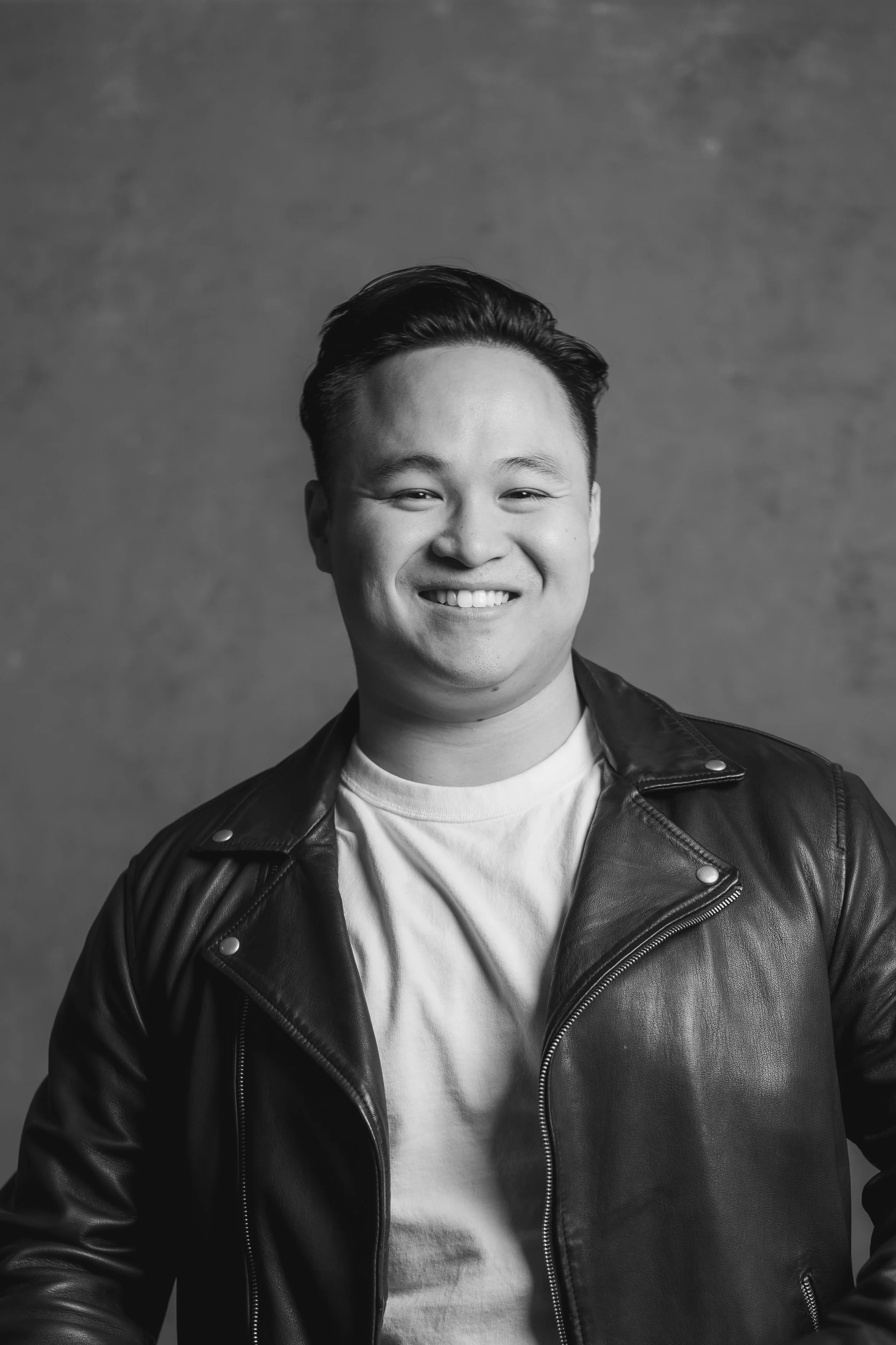 Black and white portrait of a smiling young man with short hair, wearing a leather jacket over a T-shirt, against a plain background.