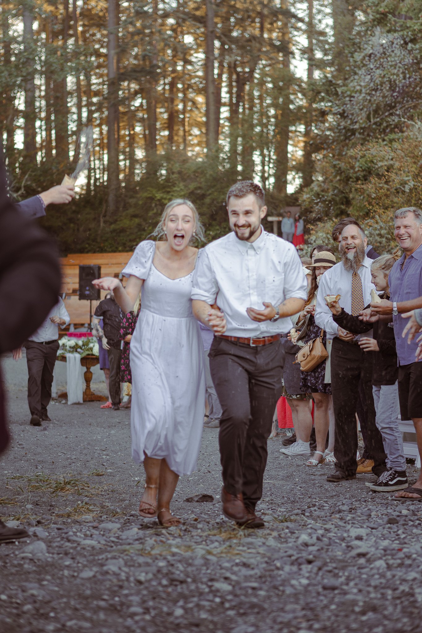 A wedding couple walking hand in hand among guests in a wooded setting.