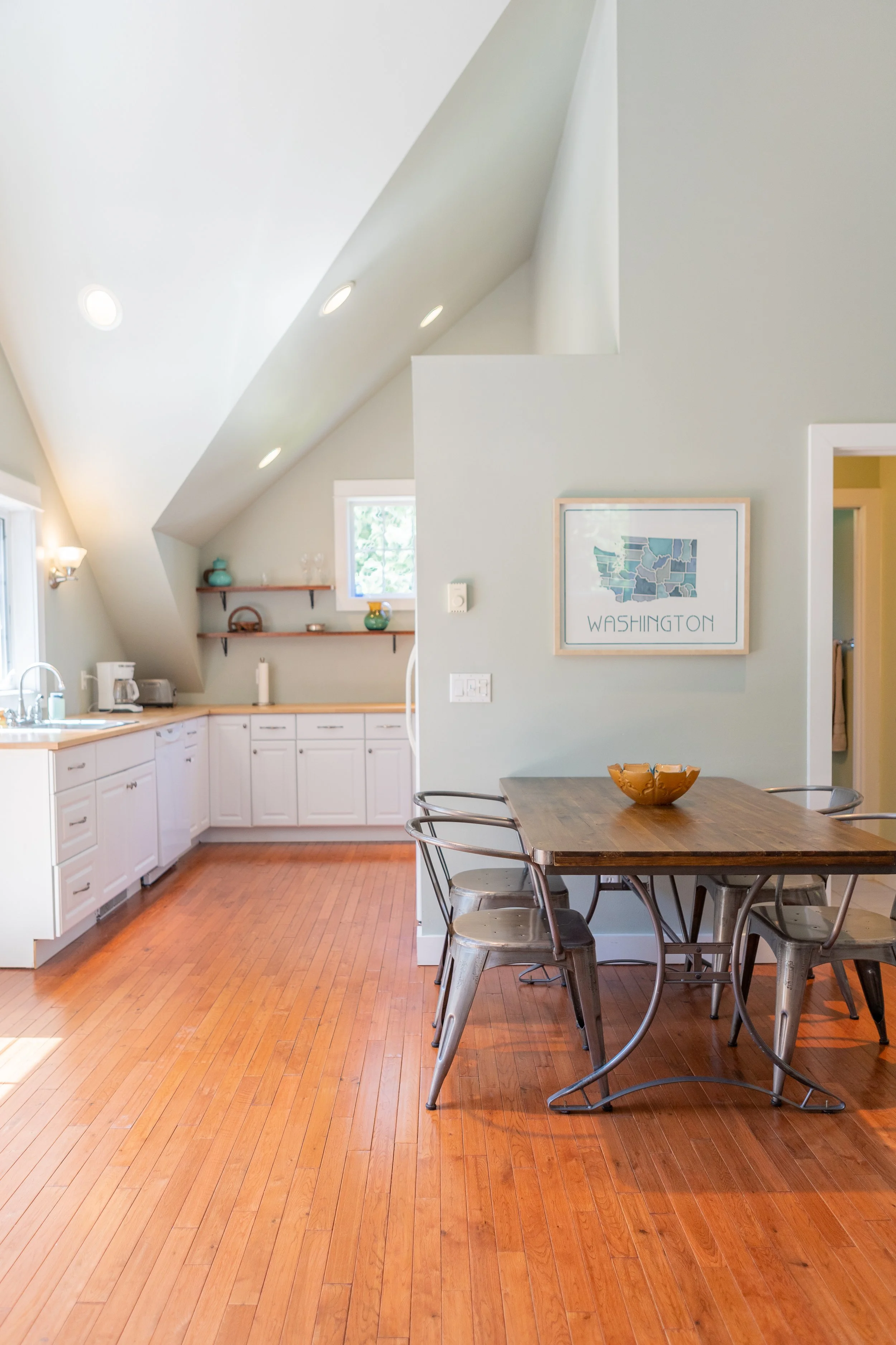 A bright kitchen with white cabinets, a wooden dining table with metal chairs, a framed map of Washington on the wall, and a sloped ceiling with recessed lighting.