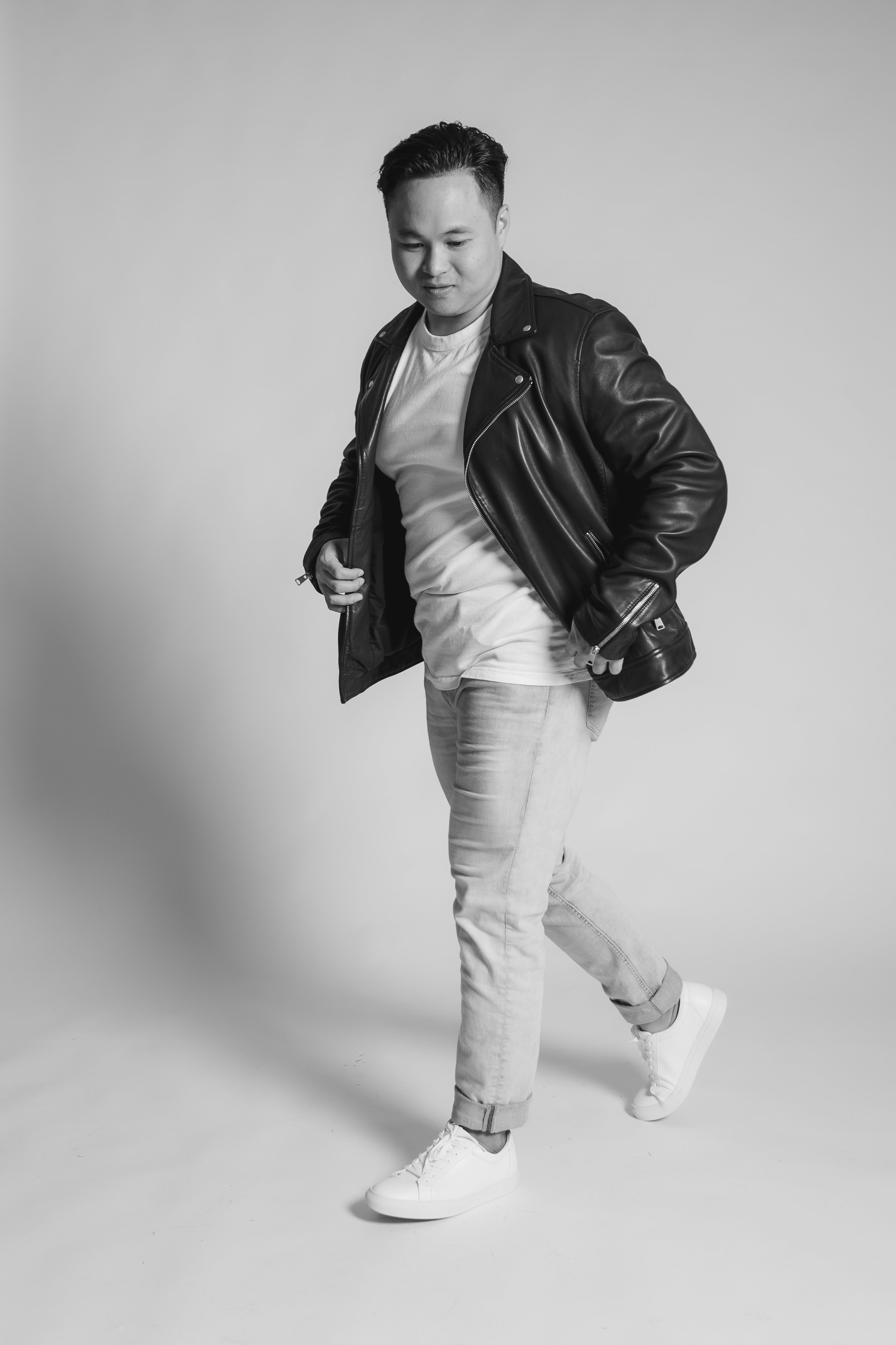 A young man dressed in a leather jacket, white T-shirt, light jeans, and white sneakers is posing with one foot raised and hands in pockets against a plain background.