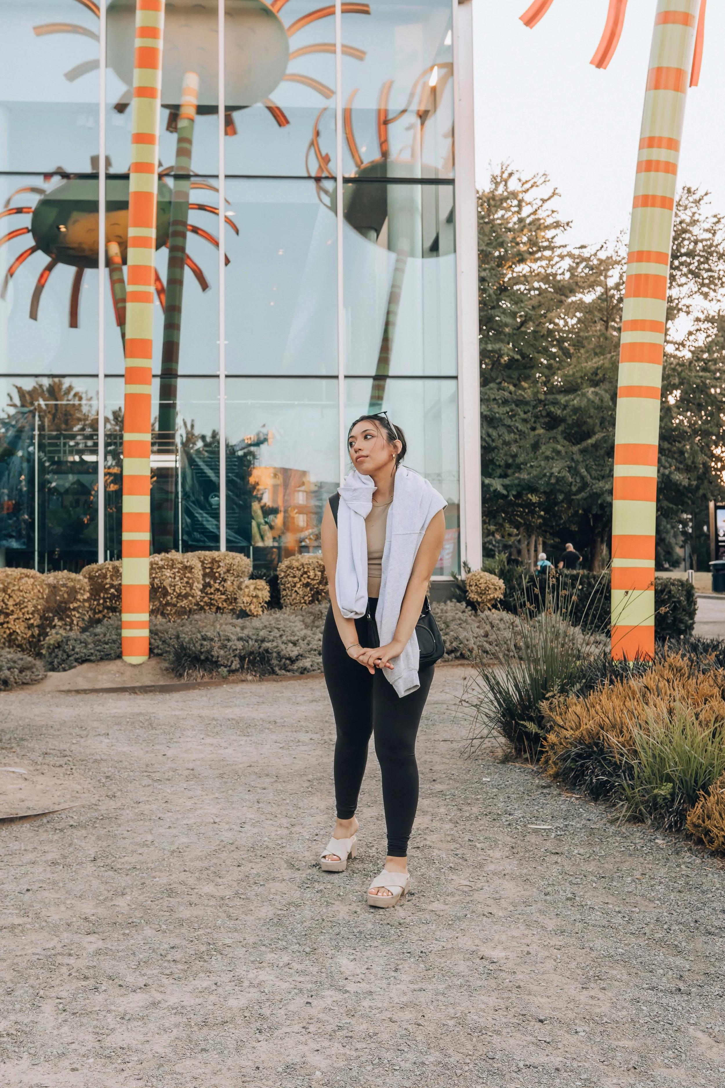 A woman with long black hair wearing a gray sleeveless top, black leggings, and white platform sandals standing outdoors in front of a modern glass building with colorful art installations resembling large striped poles and spider-like sculptures. Sh