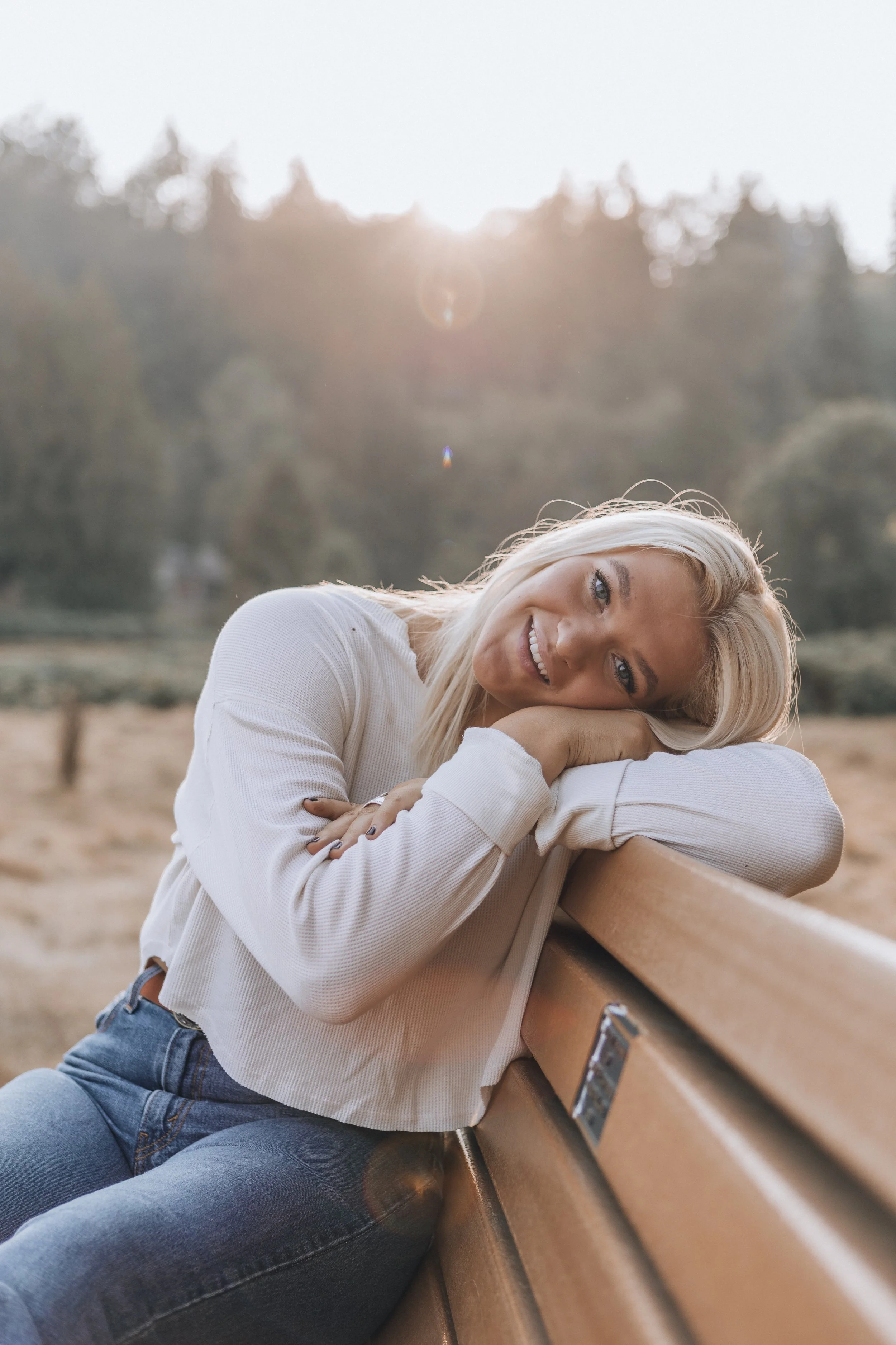 A smiling blonde woman wearing a white long sleeve top and blue jeans sitting on a park bench during sunset.
