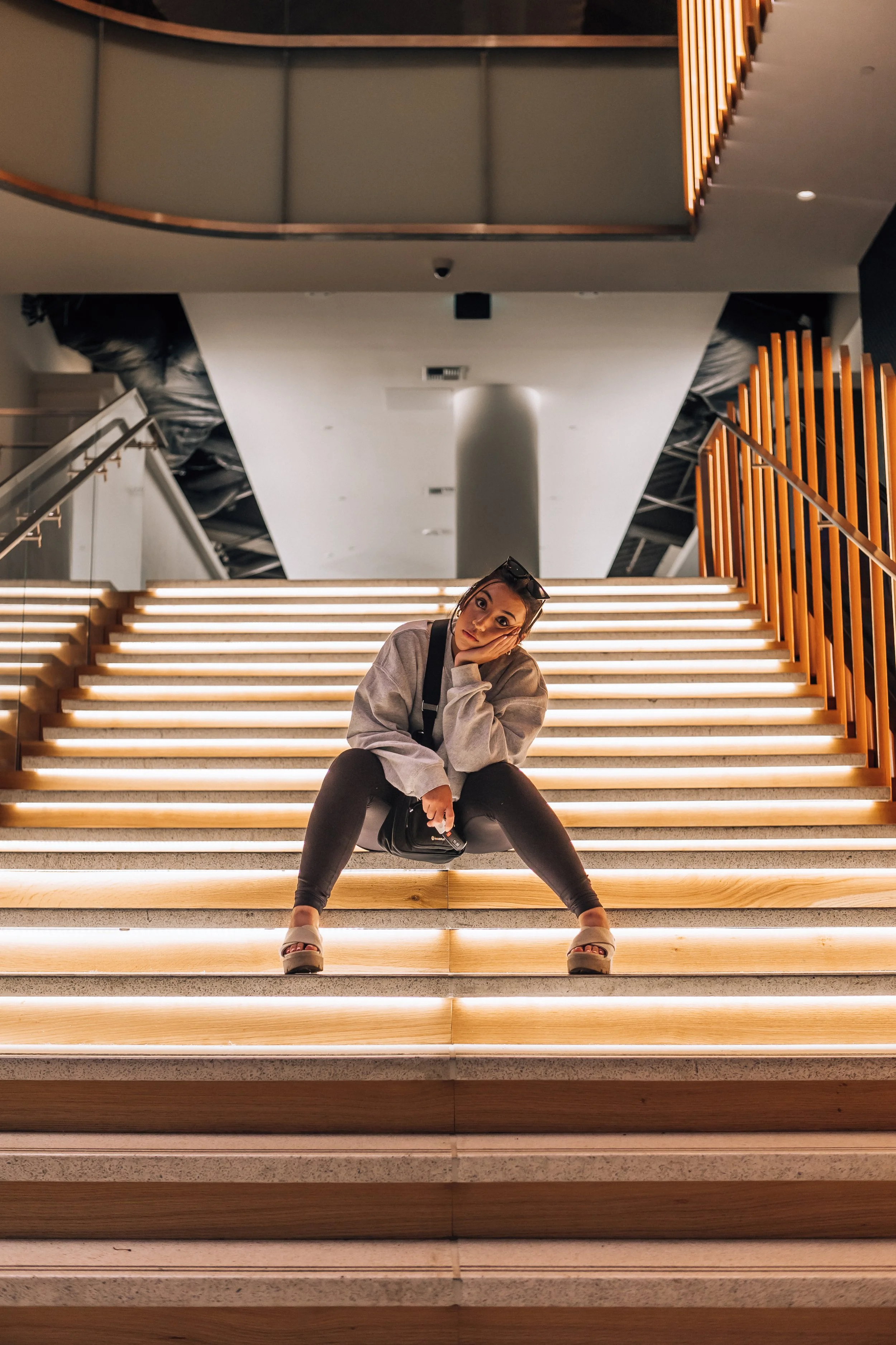 A young woman sitting on illuminated stairs inside a modern building, resting her head on her hand, with dark hair, sunglasses on her head, and wearing a gray hoodie, black pants, and white platform sandals.