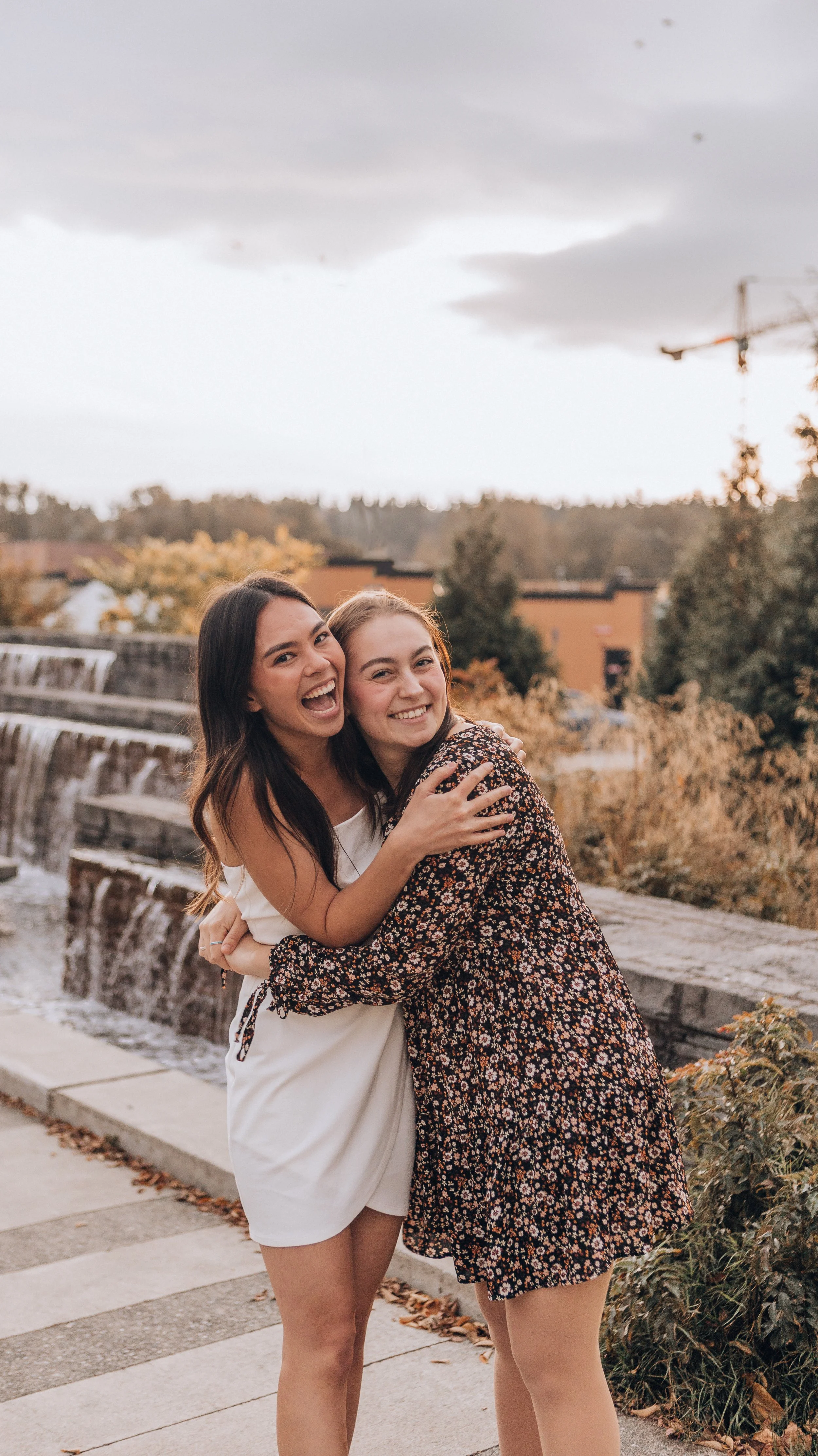 Two women hugging and smiling at the camera outdoors near a waterfall with a cloudy sky and trees in the background.