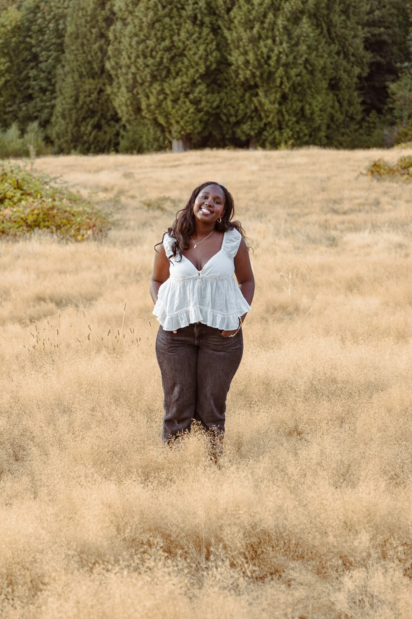 A woman standing in a field of dry grass with large trees in the background, smiling and wearing a white blouse and dark jeans.