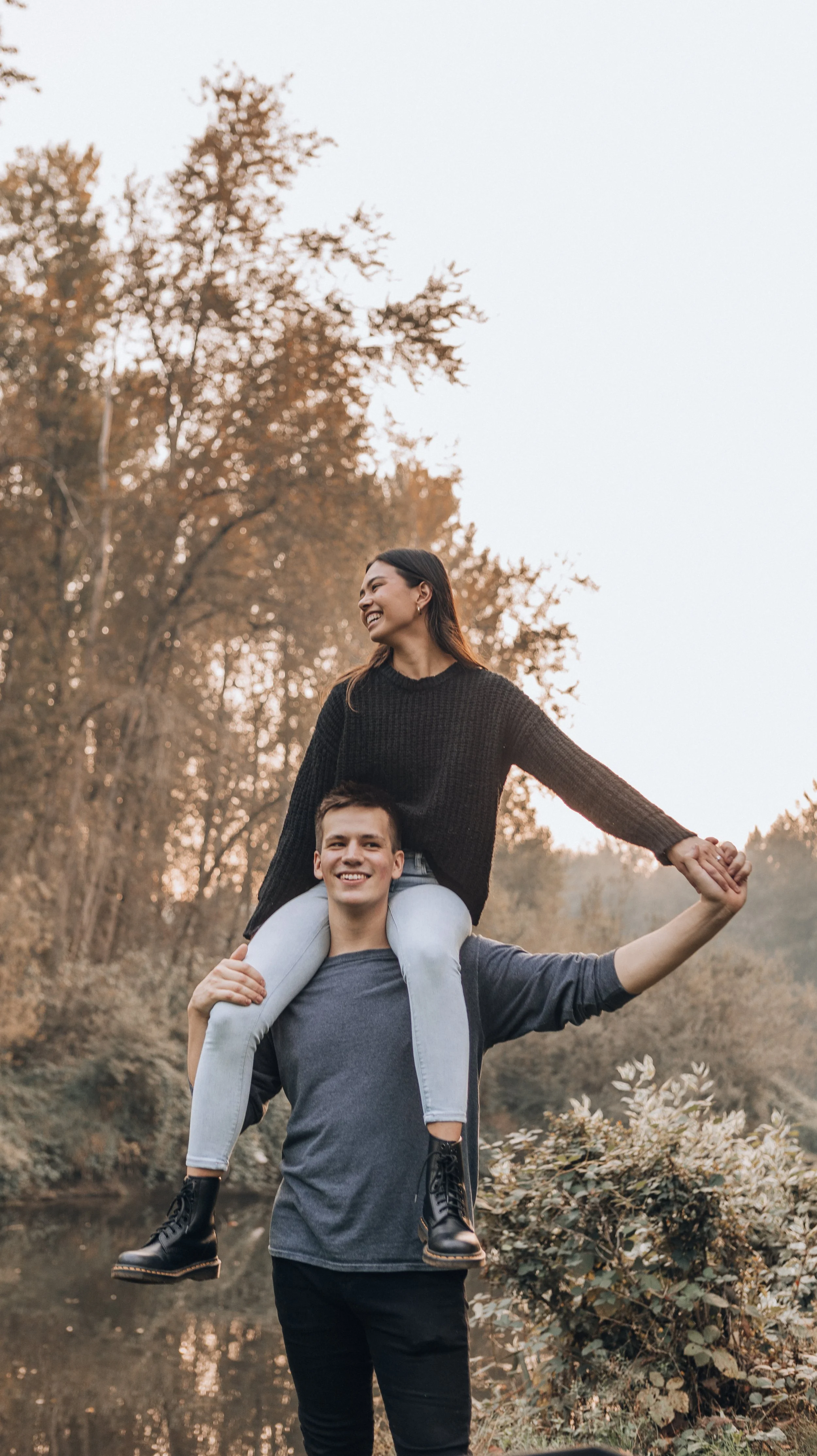 A young man is carrying a young woman on his shoulders in a park with autumn trees, both smiling and enjoying the moment.
