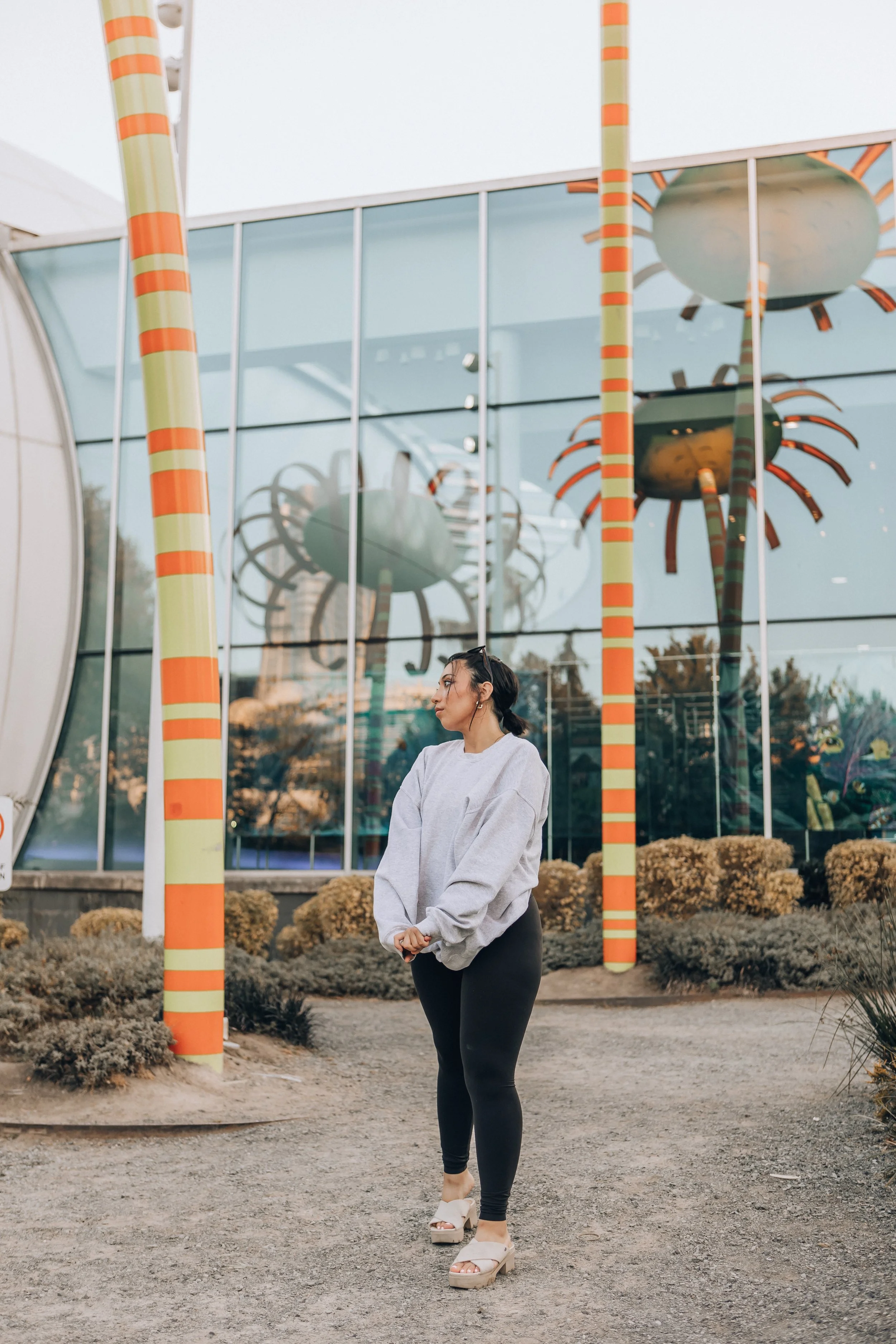 A woman standing outdoors with her arms crossed, wearing a gray sweatshirt and black leggings, in front of a modern building with reflective glass windows and colorful striped poles, during daytime.