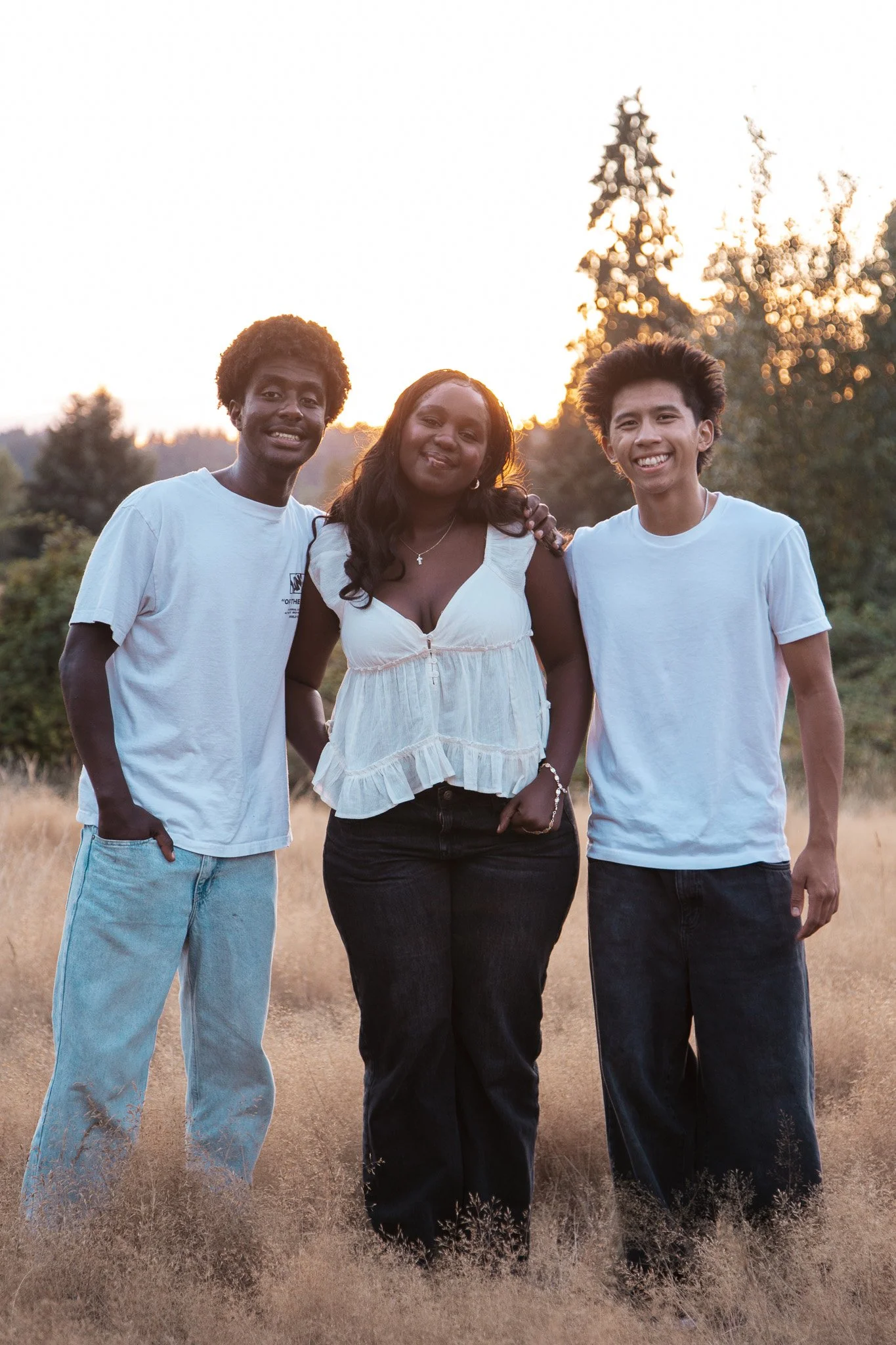 Three young people standing together outdoors during sunset, smiling at the camera. The person on the left has curly hair, wearing a white T-shirt and light blue jeans. The person in the middle has long dark hair, wearing a white sleeveless top and d