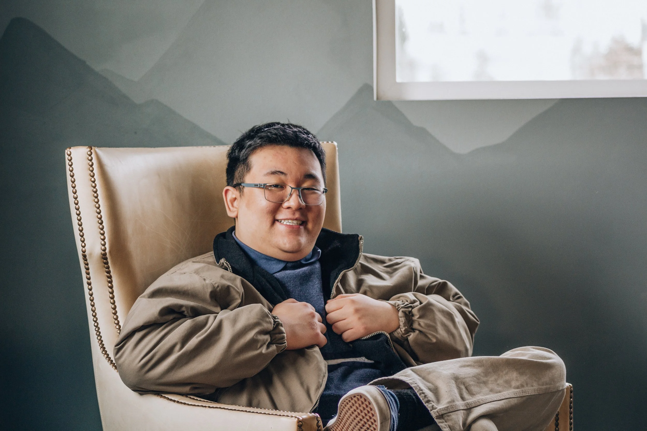 A young man with glasses smiling, sitting in a beige armchair, wearing a tan jacket, blue sweater, and jeans, in a room with a window and painted wall.