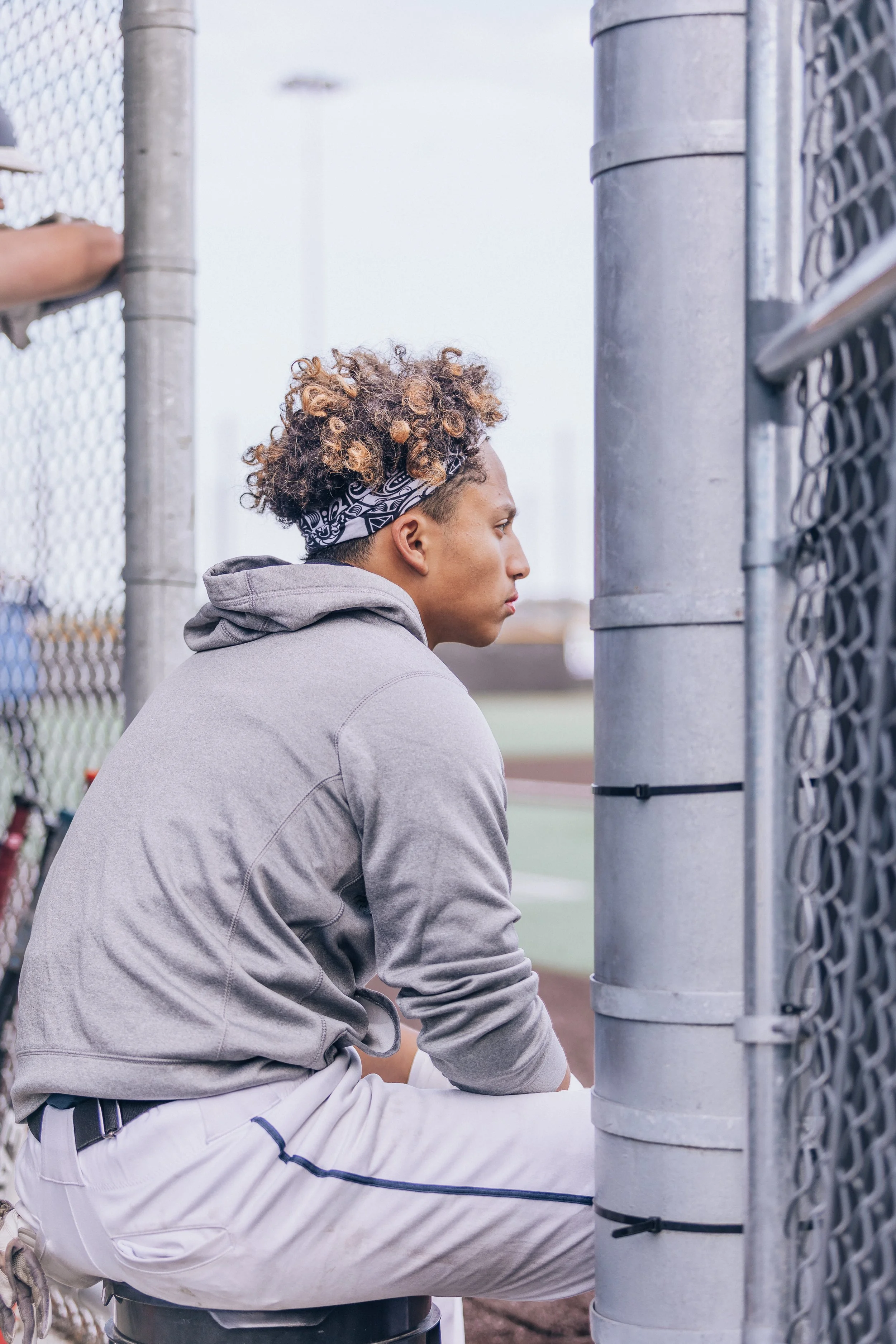 A young man with curly hair tied back with a bandana, wearing a gray hoodie and white athletic pants, sitting on a stool at a fenced outdoor sports court.