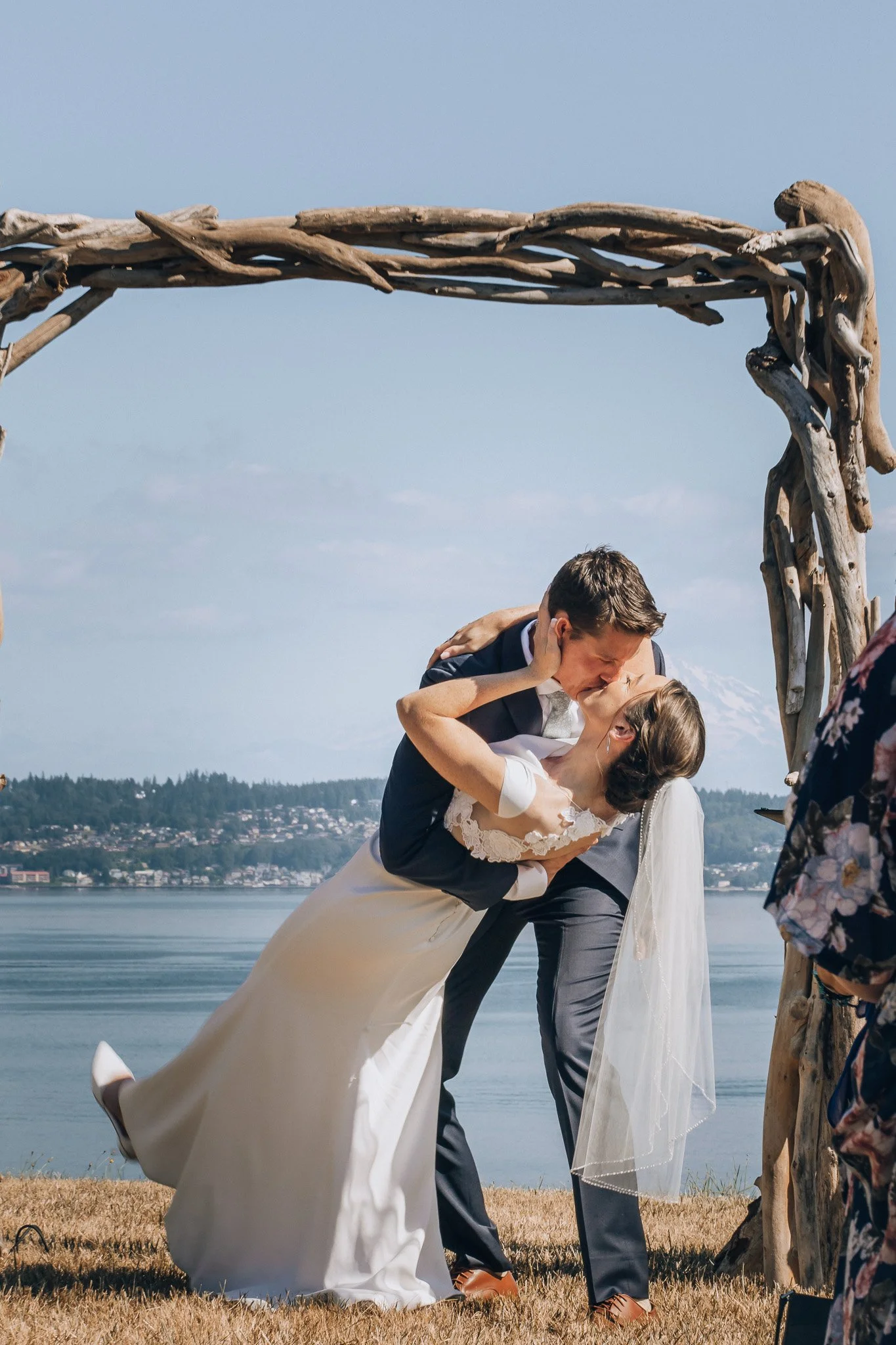A wedding couple beside a driftwood arch overlooking the water and mountains.