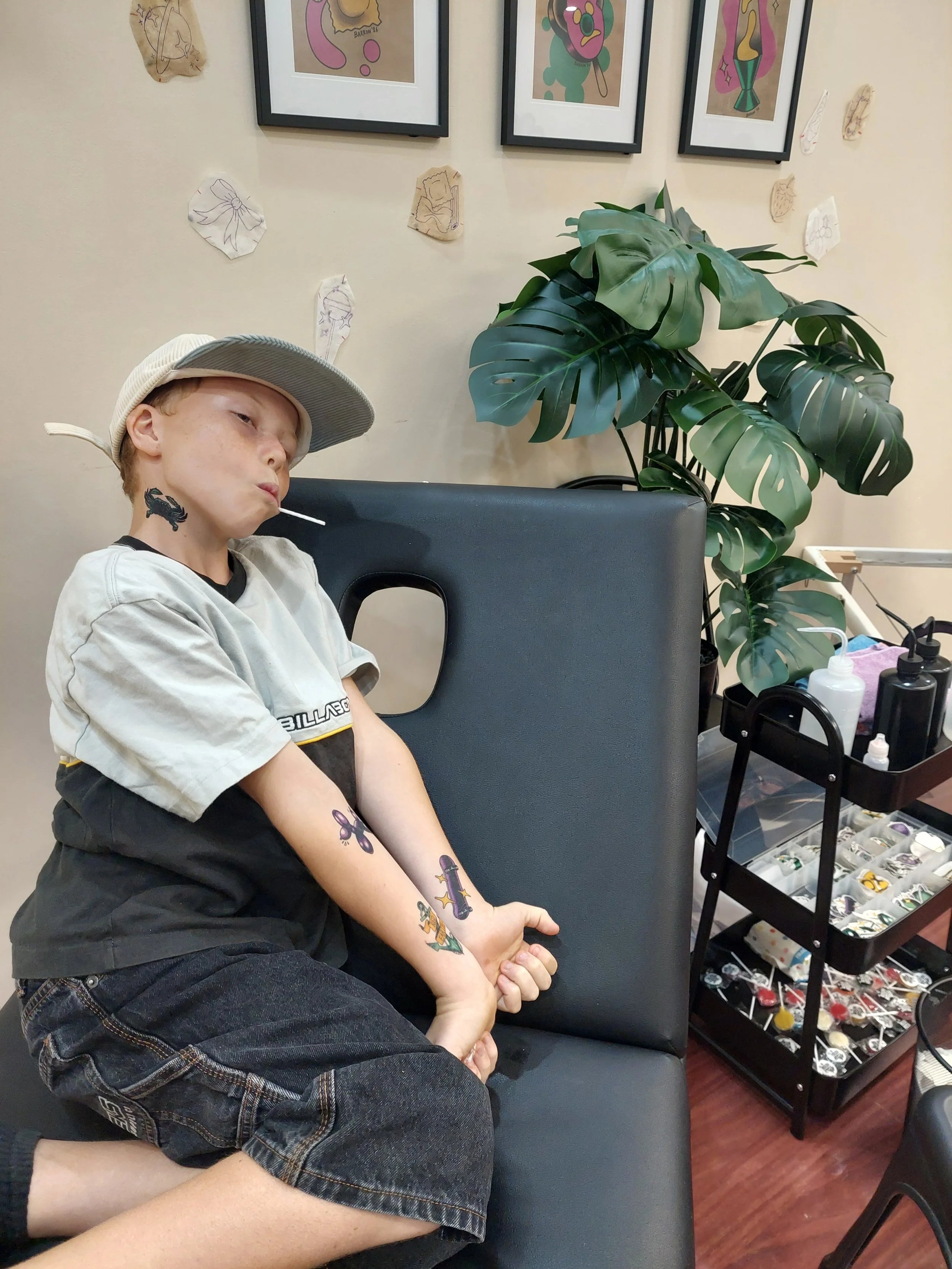 A young boy poses with a lollipop in his mouth and both arms extended to show off his many temporary tattoos
