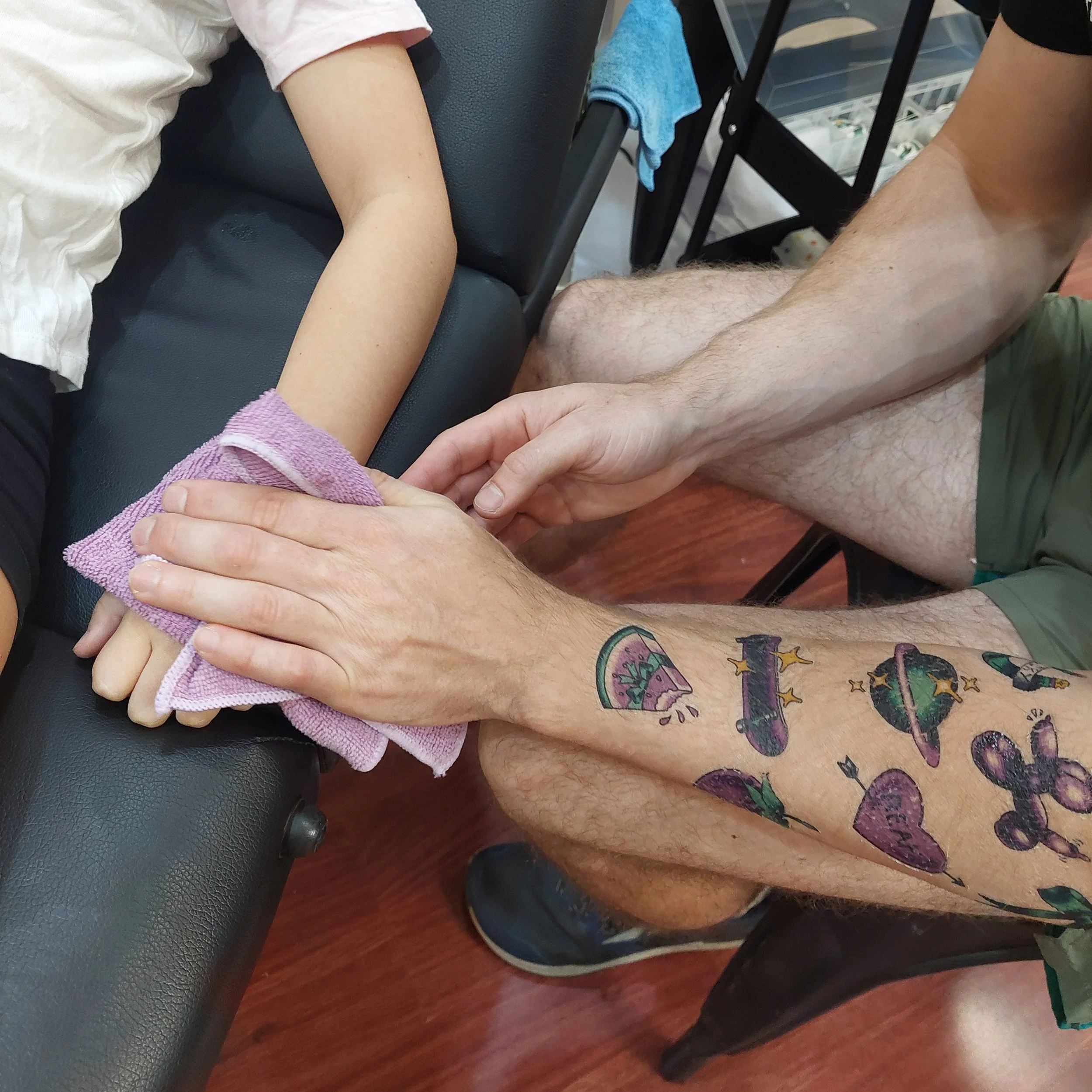 An arm adorned with a number of temporary tattoos presses a cloth onto a small childs hand in the process of applying a temporary tattoo