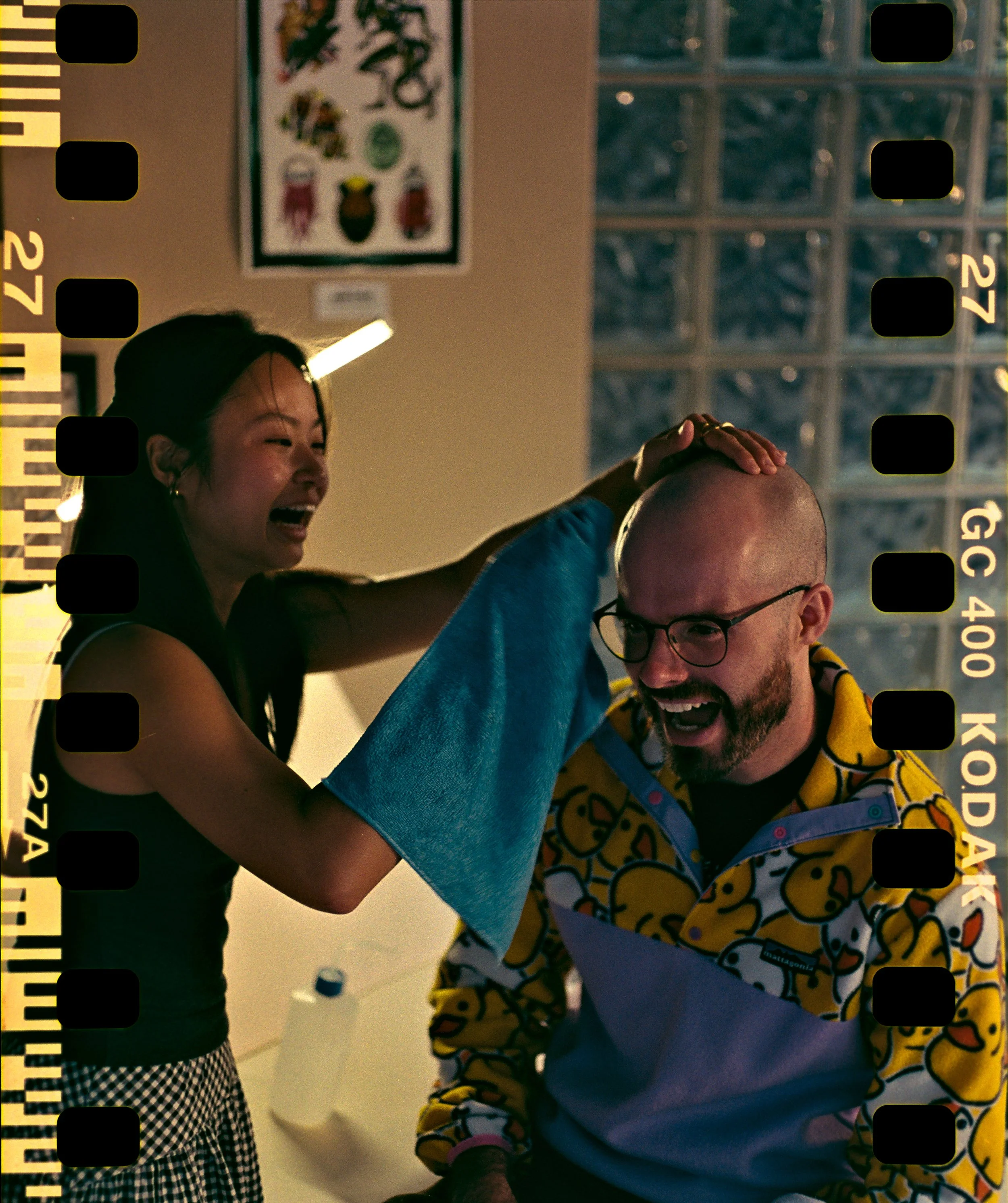 A performer laughs as she applies a temporary tattoo to a smiling bald man's head
