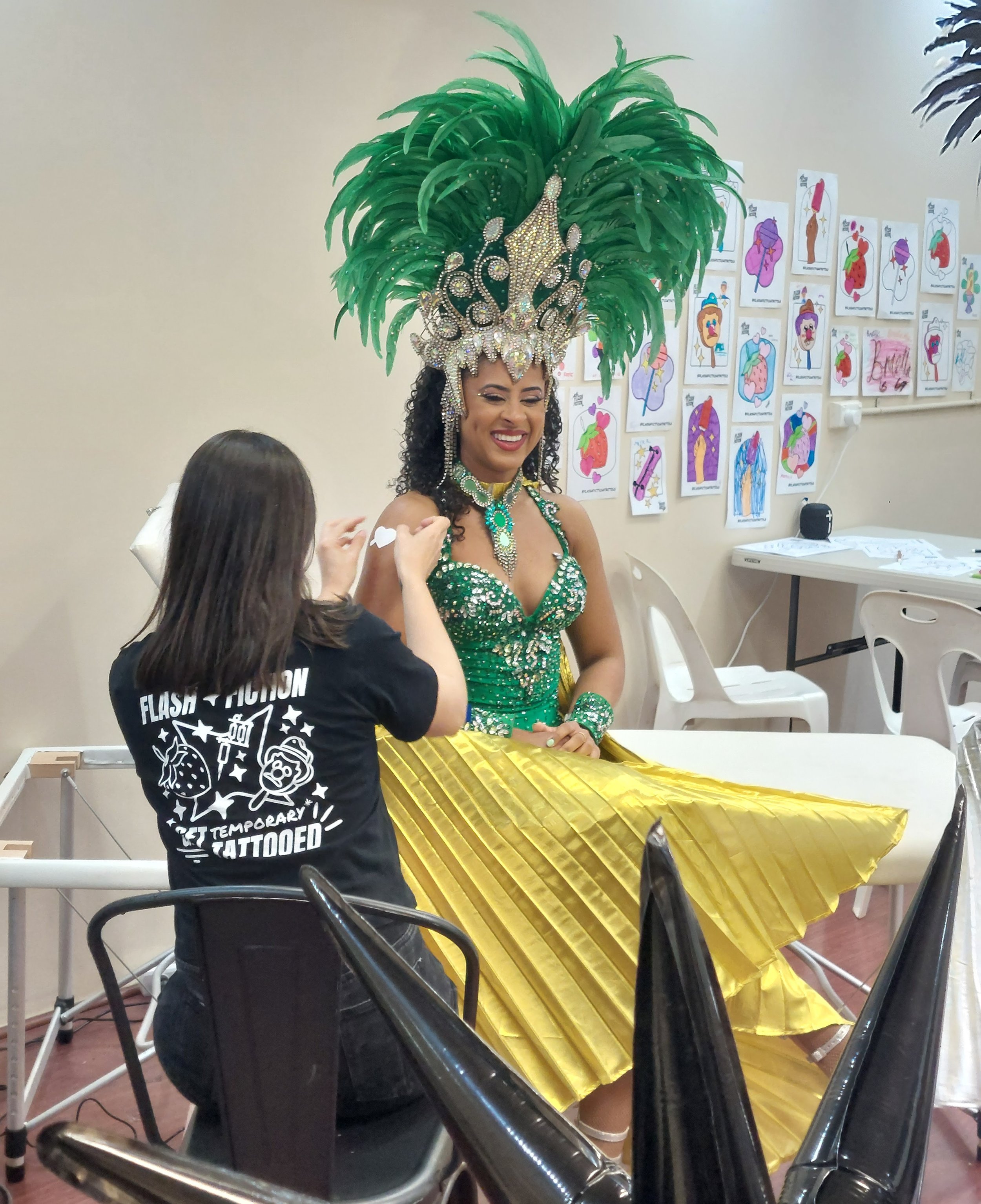 An extravagantly dressed dance performer in a feathered headress smiles as a performer applies a temporary tattoo to her shoulder. The dancer is seated on a tattoo bed with tattoo art on the walls behind her
