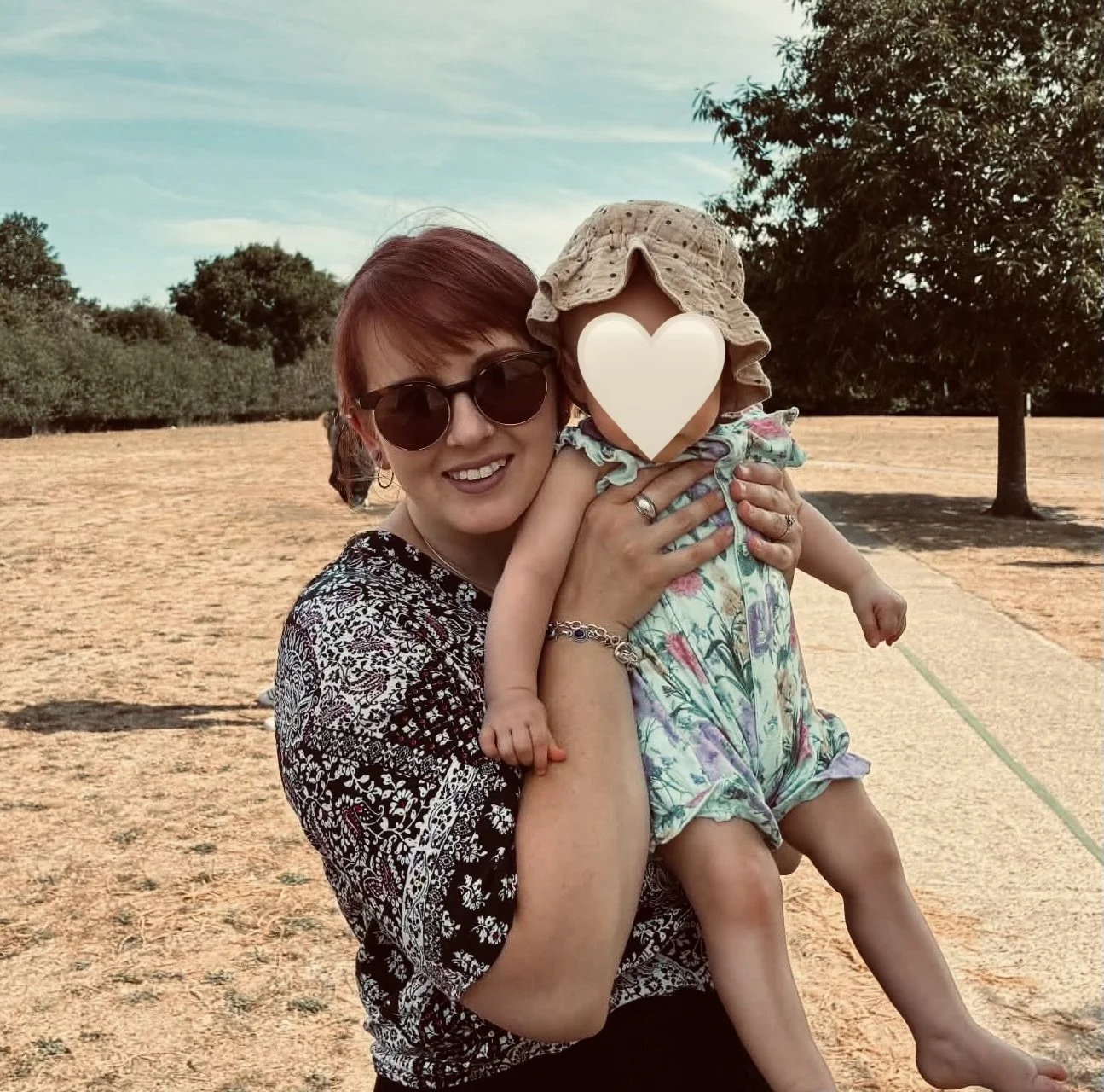 Woman holding a young child outdoors on a sunny day, with trees and a grassy field in the background.
