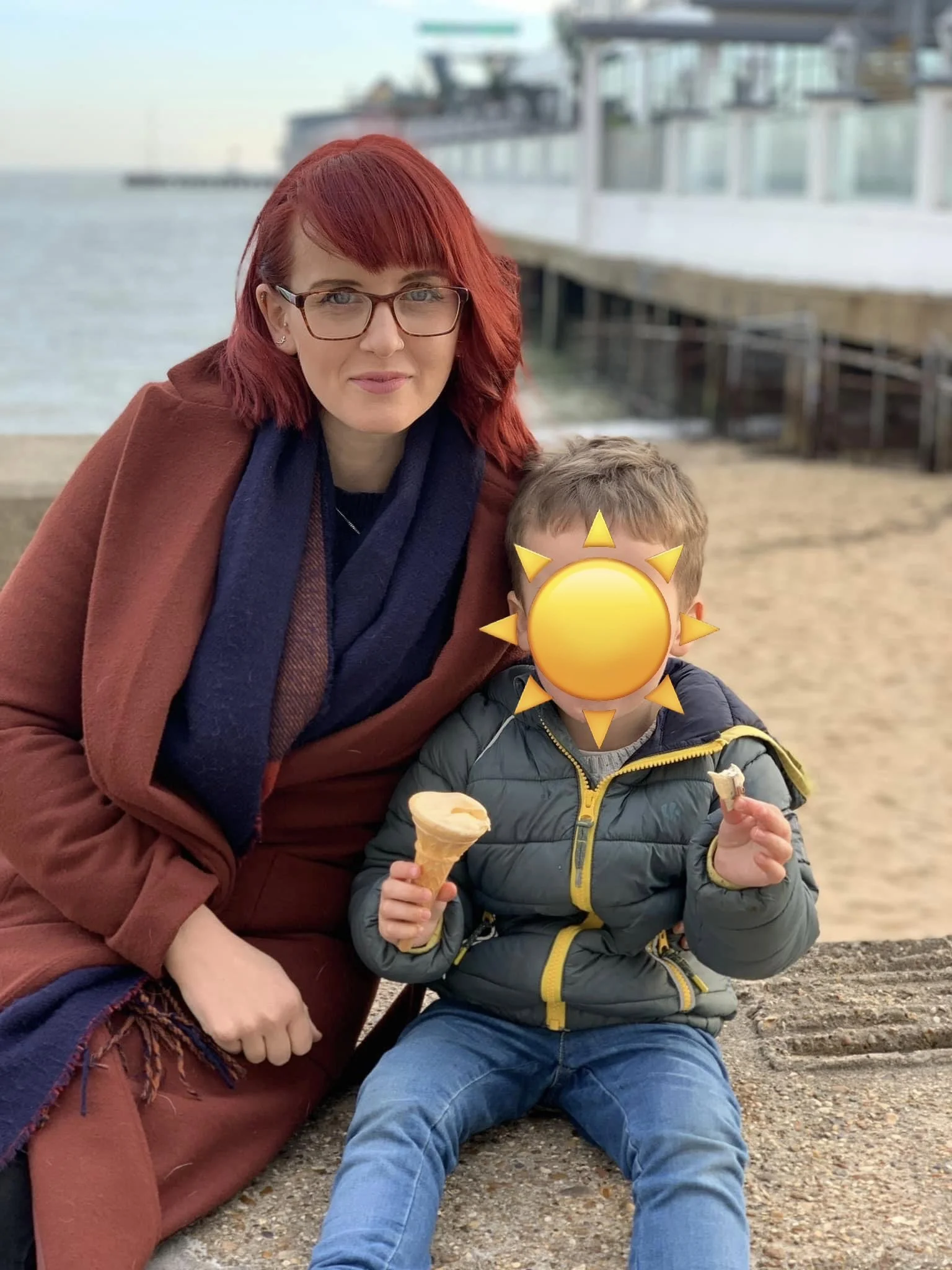 A woman with red hair, glasses, and a brown coat sitting on the ground by a child holding ice cream cone at the beach near a pier and water.
