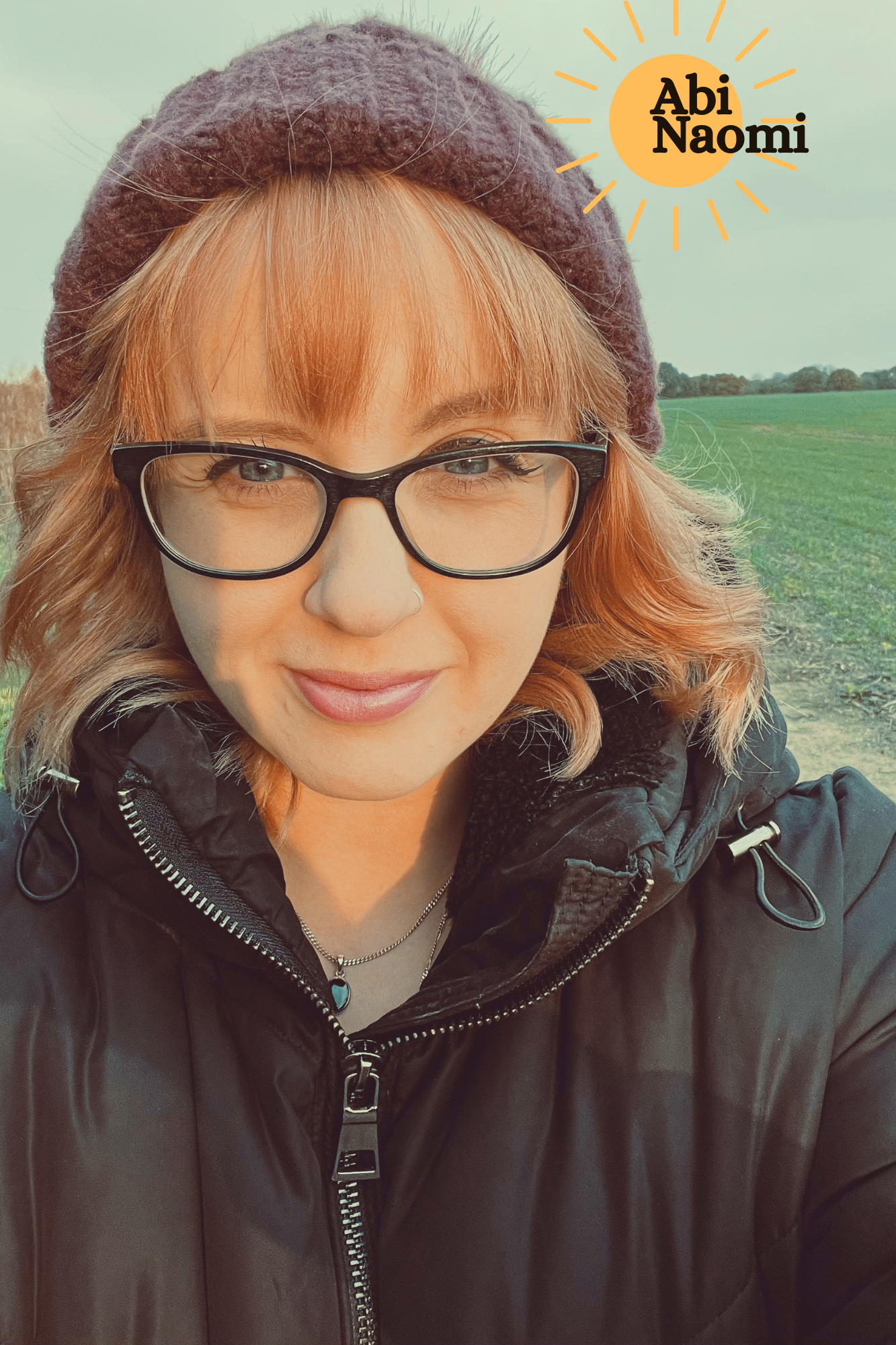 Close-up of a person with glasses, orange hair, wearing a beanie and a black jacket, outdoors in a wide open field. The photo includes a graphic of a sun with the text "Abi Naomi" in it.