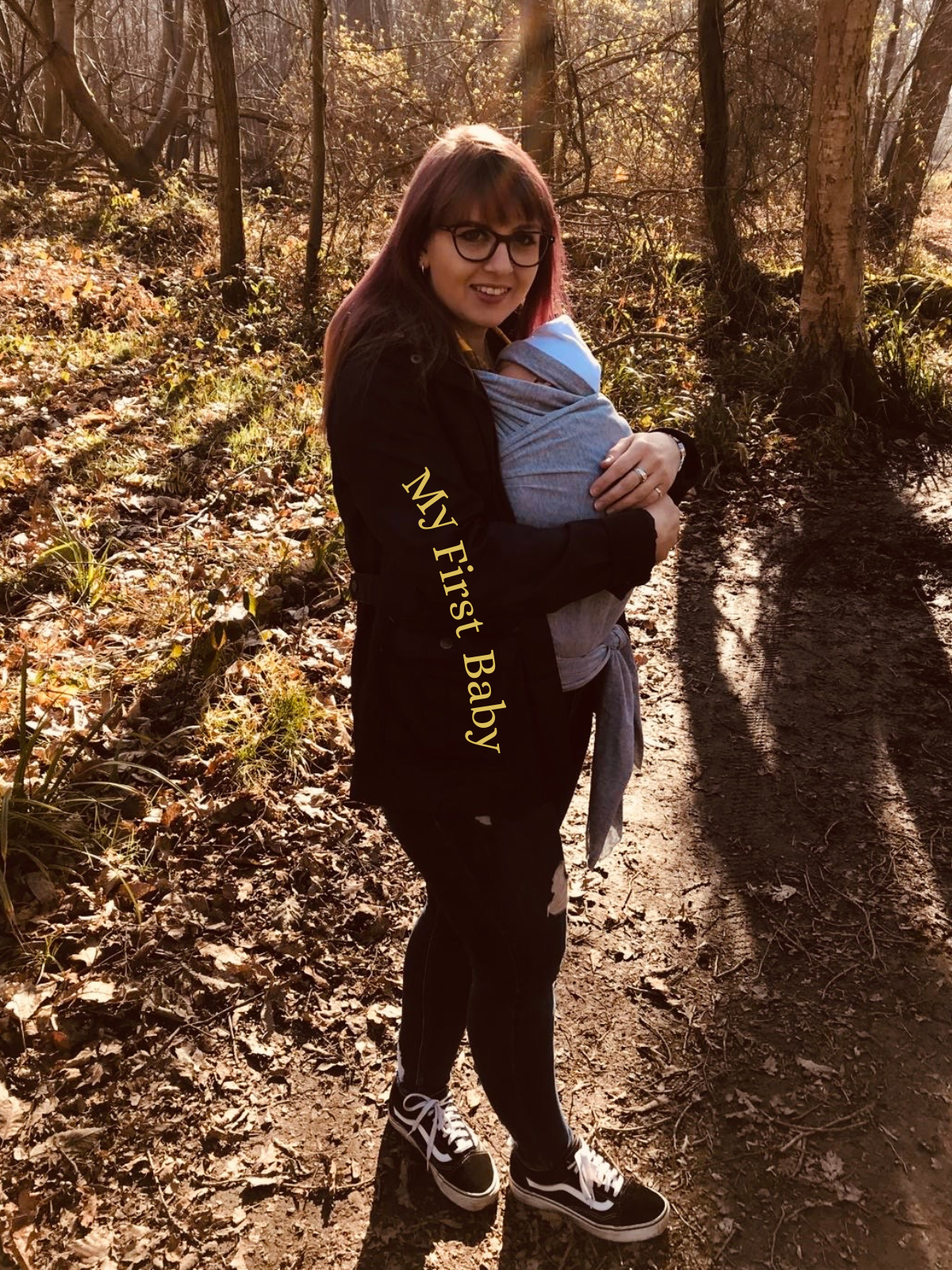 A woman in glasses and casual clothing holding a baby in a gray carriersack on a dirt trail in a sunlit wooded area. The woman is smiling at the camera, and the text overlay reads "My First Baby."