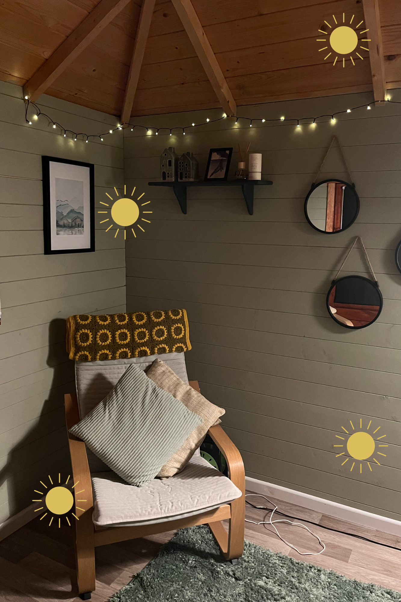 A cozy corner of a room with a wooden armchair, pillows, a knitted throw blanket, wall art, a shelf, and hanging round mirrors, decorated with string lights and a rug on the floor.