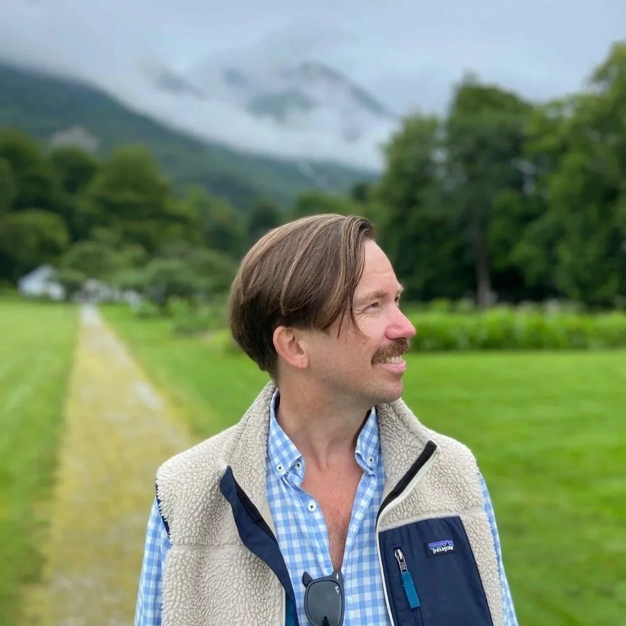 En mann med brunt hår og skjegg smiler og ser til høyre. Han står på en grønn sti i et landskap med trær og fjell i bakgrunnen.