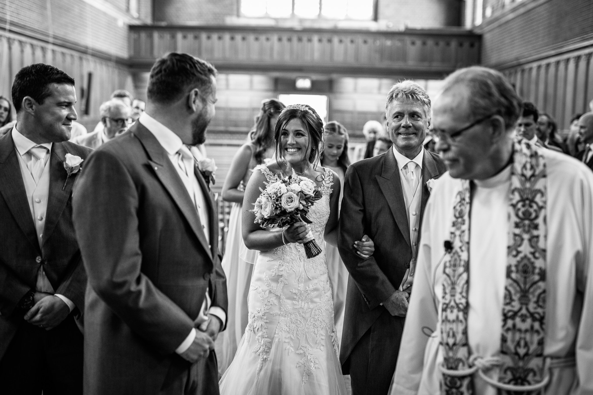  Bride smiles at the Groom as they meet at the top of the aisle  at the Chapel of St Luke at Epsom College 