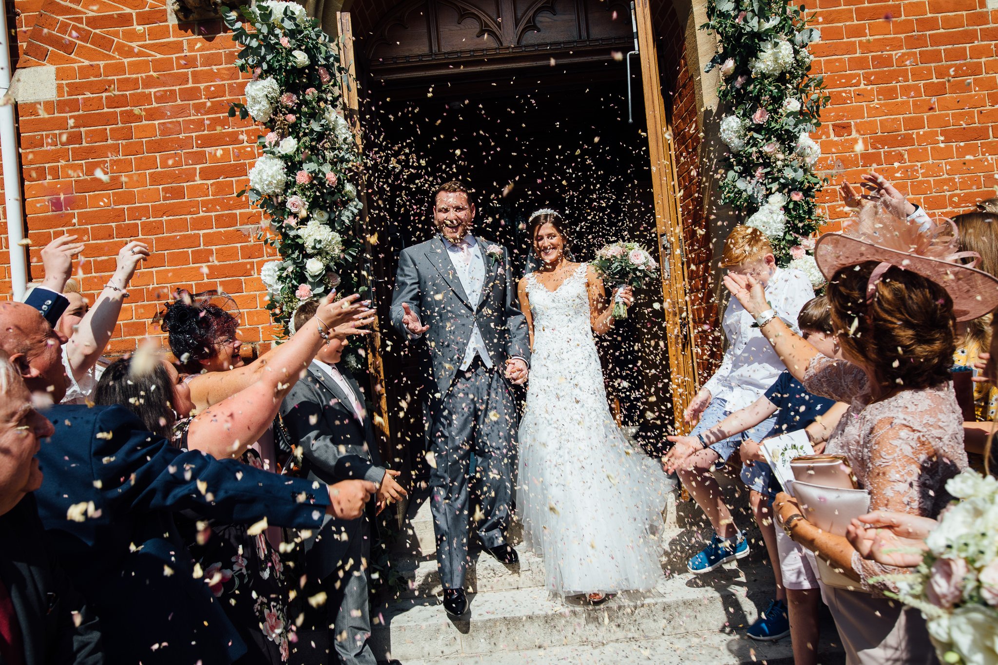  Bride and groom have confetti them at them outside  the Chapel of St Luke at Epsom College 