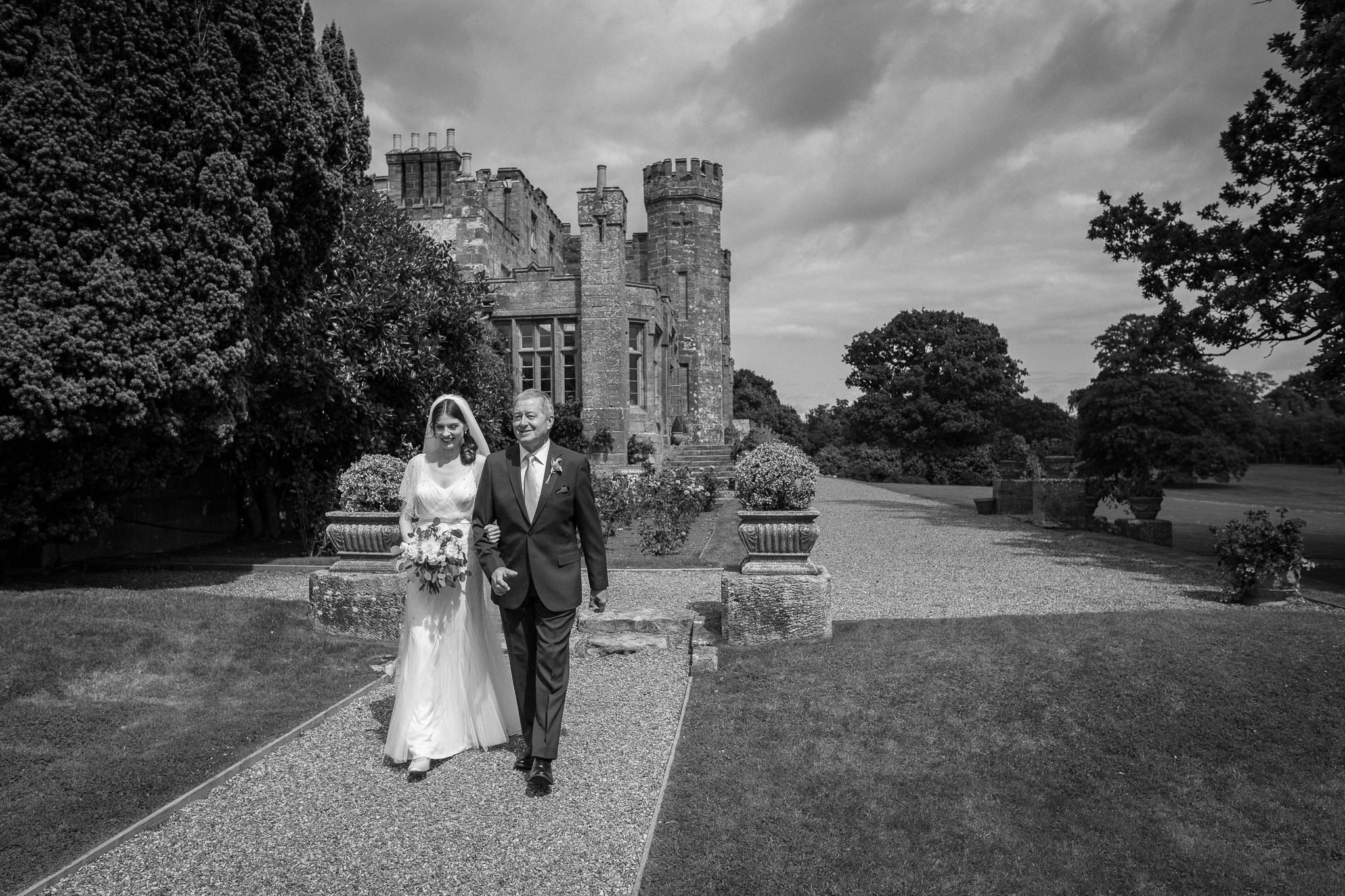  Bride and her father walk to the ceremony with  Wadhurst castle wedding venue building in the background 
