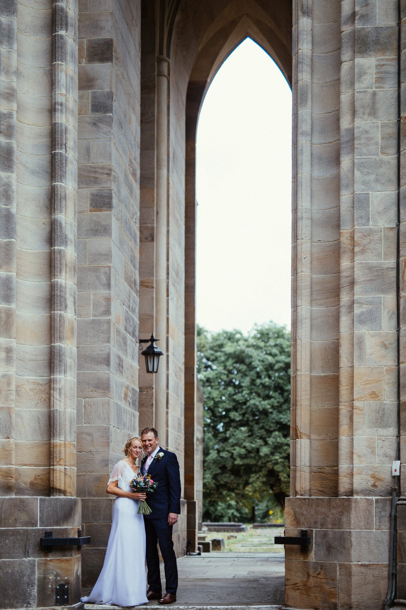  Bride and groom stand under an archway outside of All Saints Stand Church Whitefield 