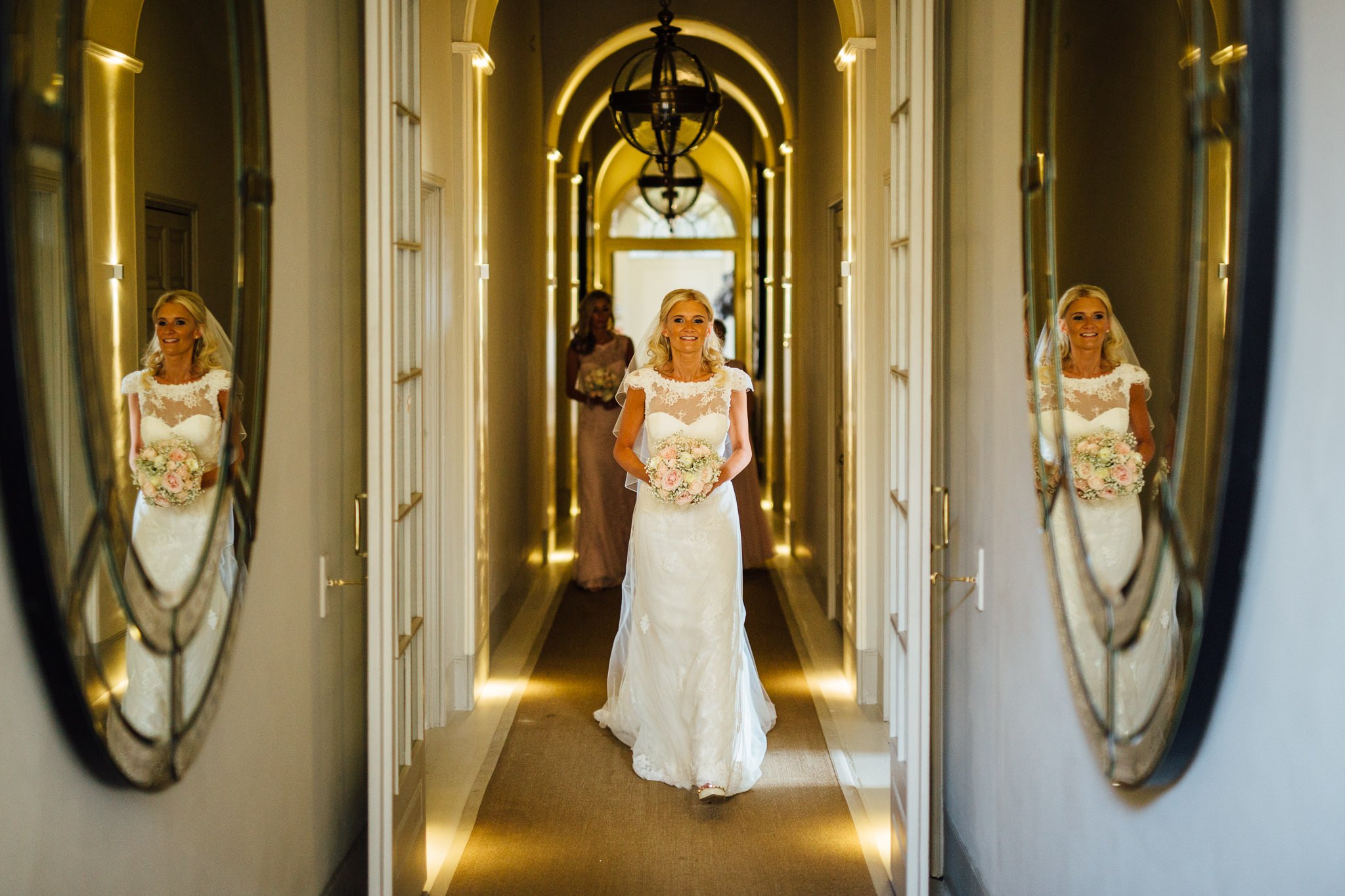  Bride walks to the wedding ceremony down the corridors of Aynhoe Park flanked by mirrors on the wall 