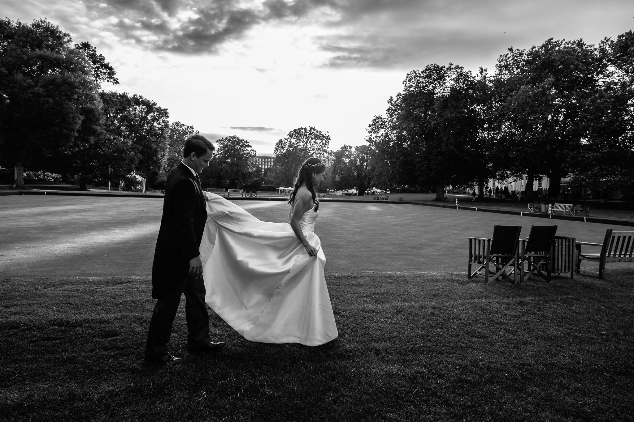  Groom carries the Bride’s dress train through the gardens of The Hurlingham Club  