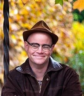 A Creatve Space member with glasses wearing a brown fedora and dark jacket, smiling outdoors with autumn leaves in the background.