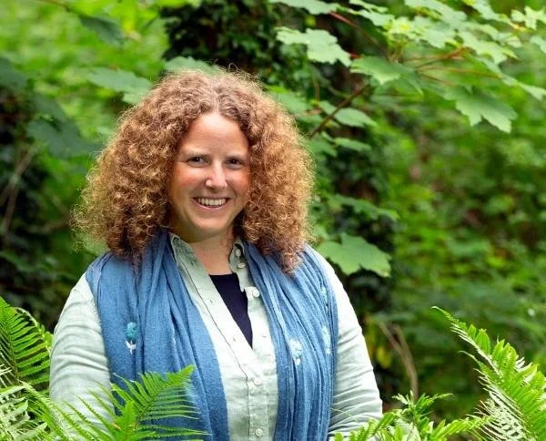 A woman with curly hair, wearing a light green shirt and patterned pants, sitting on a large rock outdoors and laughing.