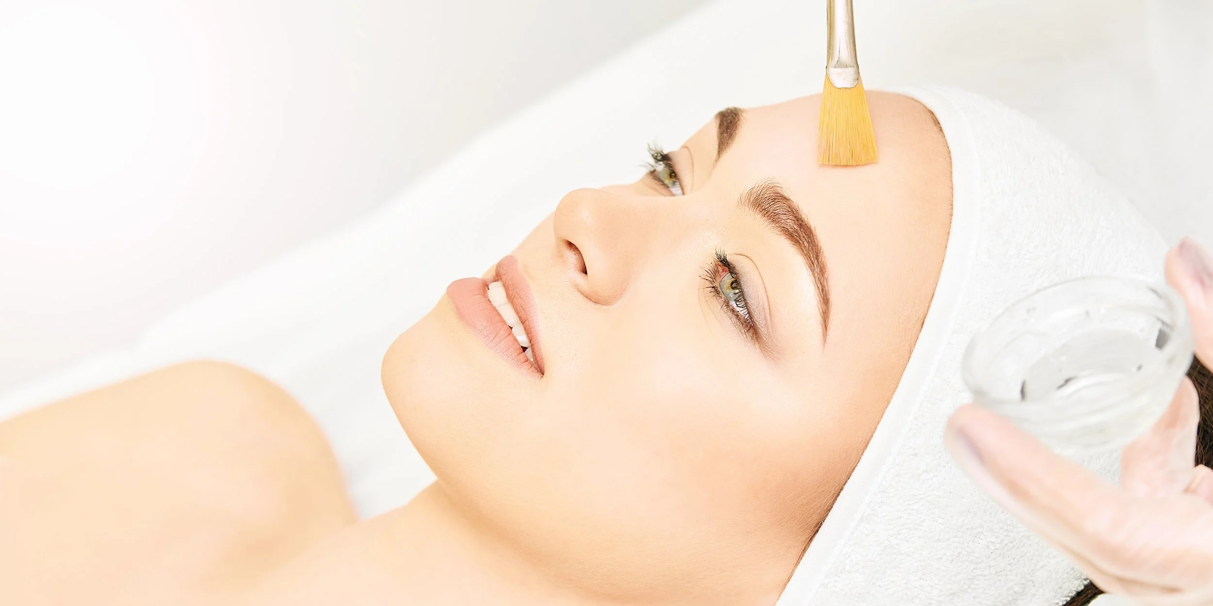 A woman receiving a beauty regenerative cosmetic treatment with a brush applying product on her forehead, lying down with a towel on her head.