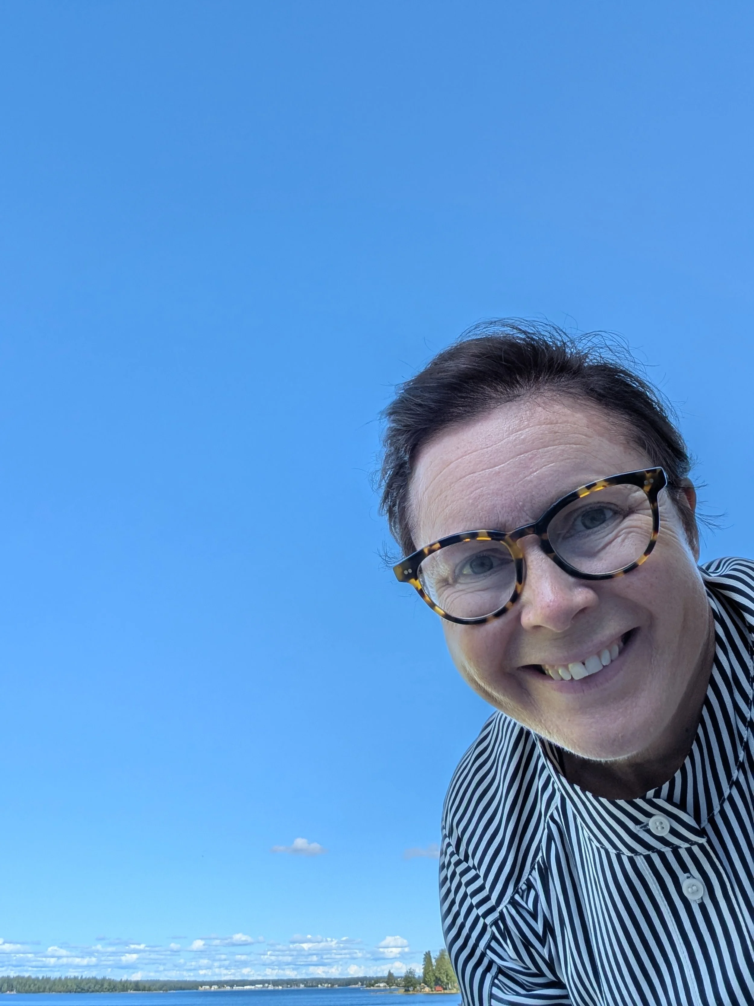 En person med mörkt hår, glasögon och randig skjorta, vänder sig mot kameran och ler mot en klar blå himmel med några moln i bakgrunden, nära en vattendrag.