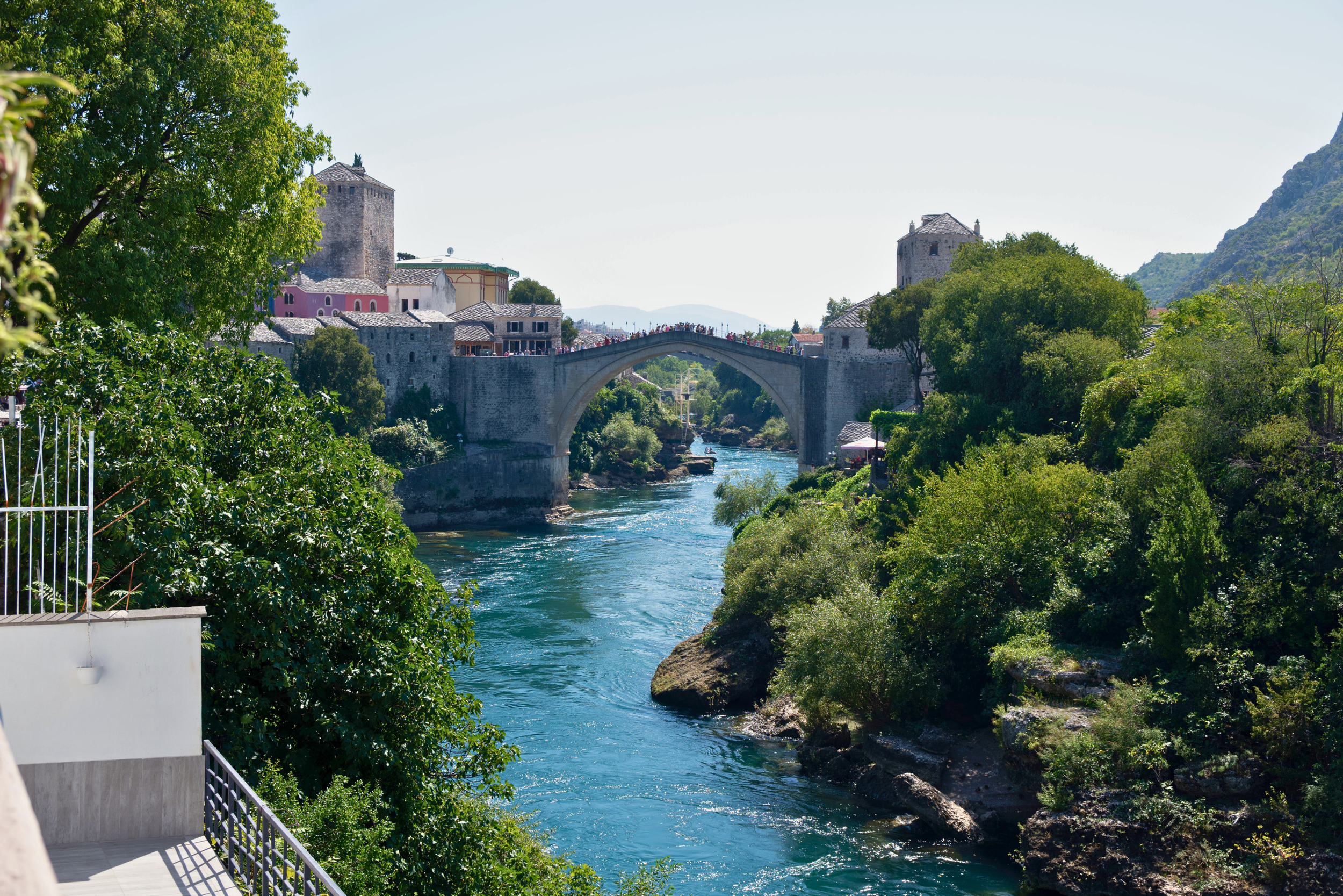 A scenic view of a historic stone bridge over a river, surrounded by lush green trees and old buildings on both sides.