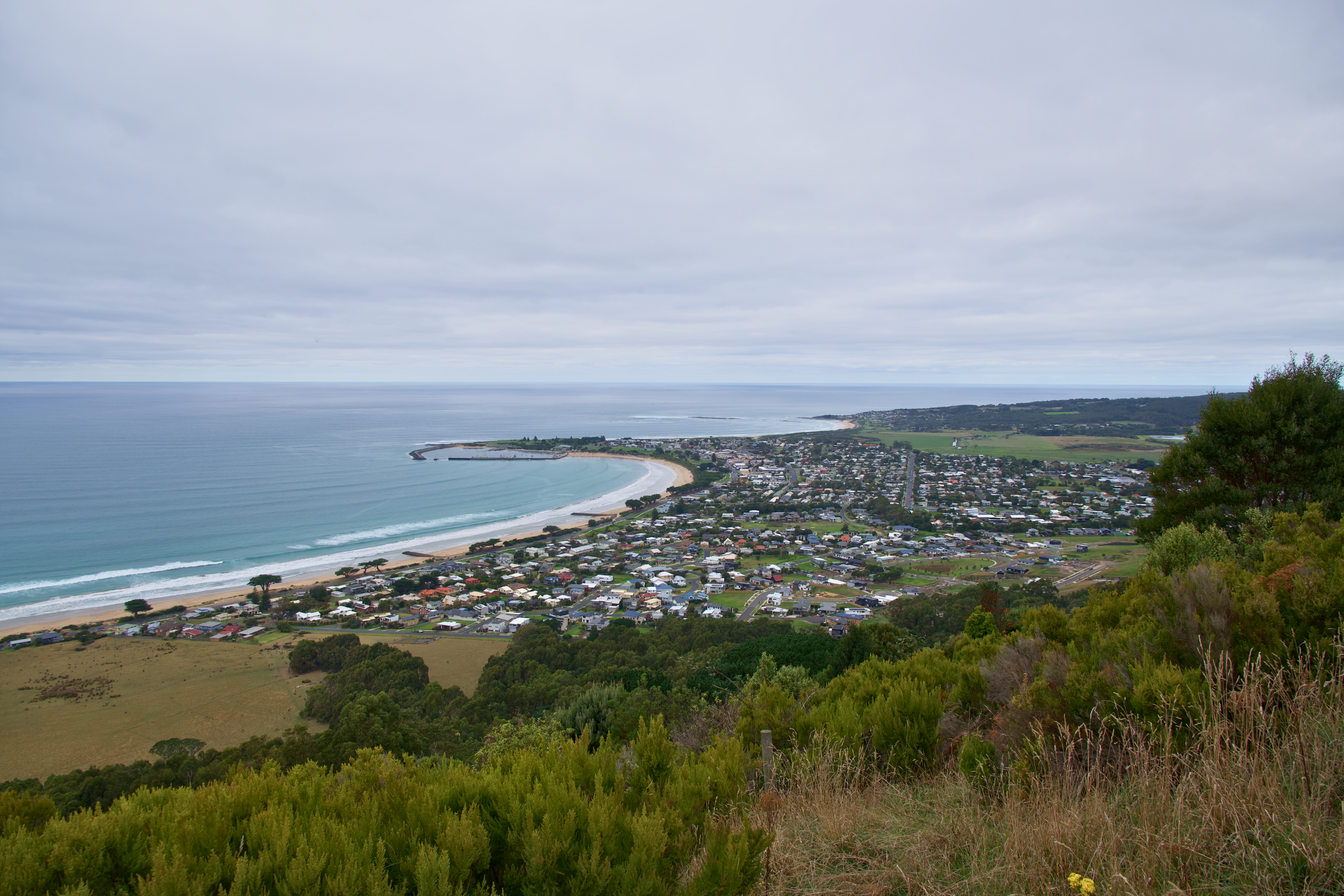 A coastal town viewed from a hillside with green shrubbery in the foreground. The town has numerous houses along the beach, with a curved shoreline and waves in the ocean. The sky is overcast with gray clouds.
